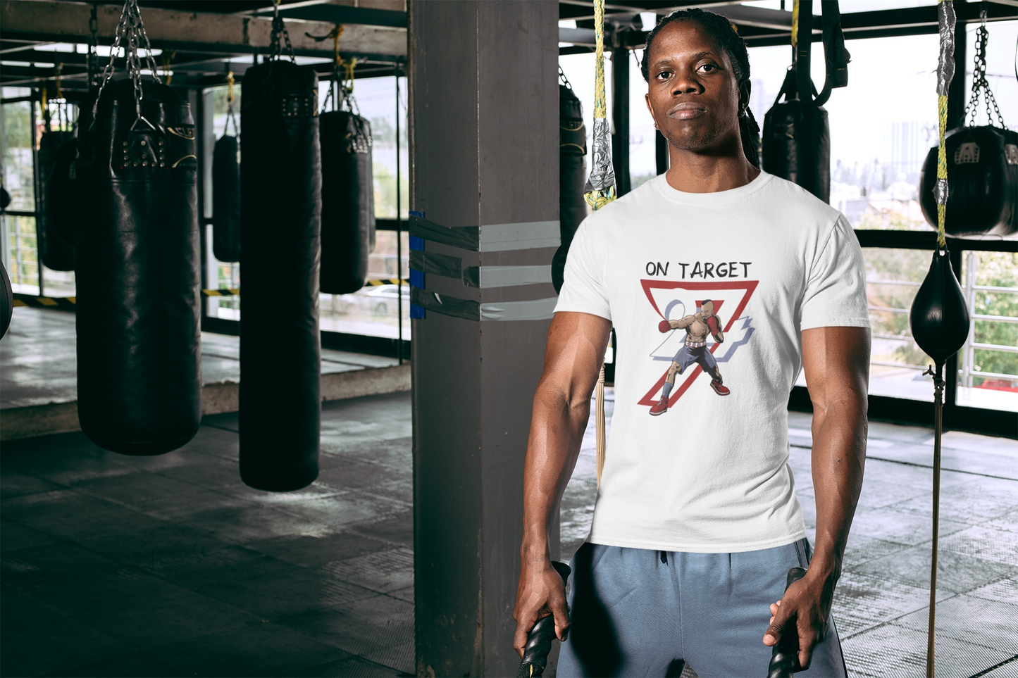 Serious male boxer standing confidently in an industrial boxing gym next to heavy bags, wearing the crisp white hand-drawn sports event shirt to showcase its structured streetwear fit.