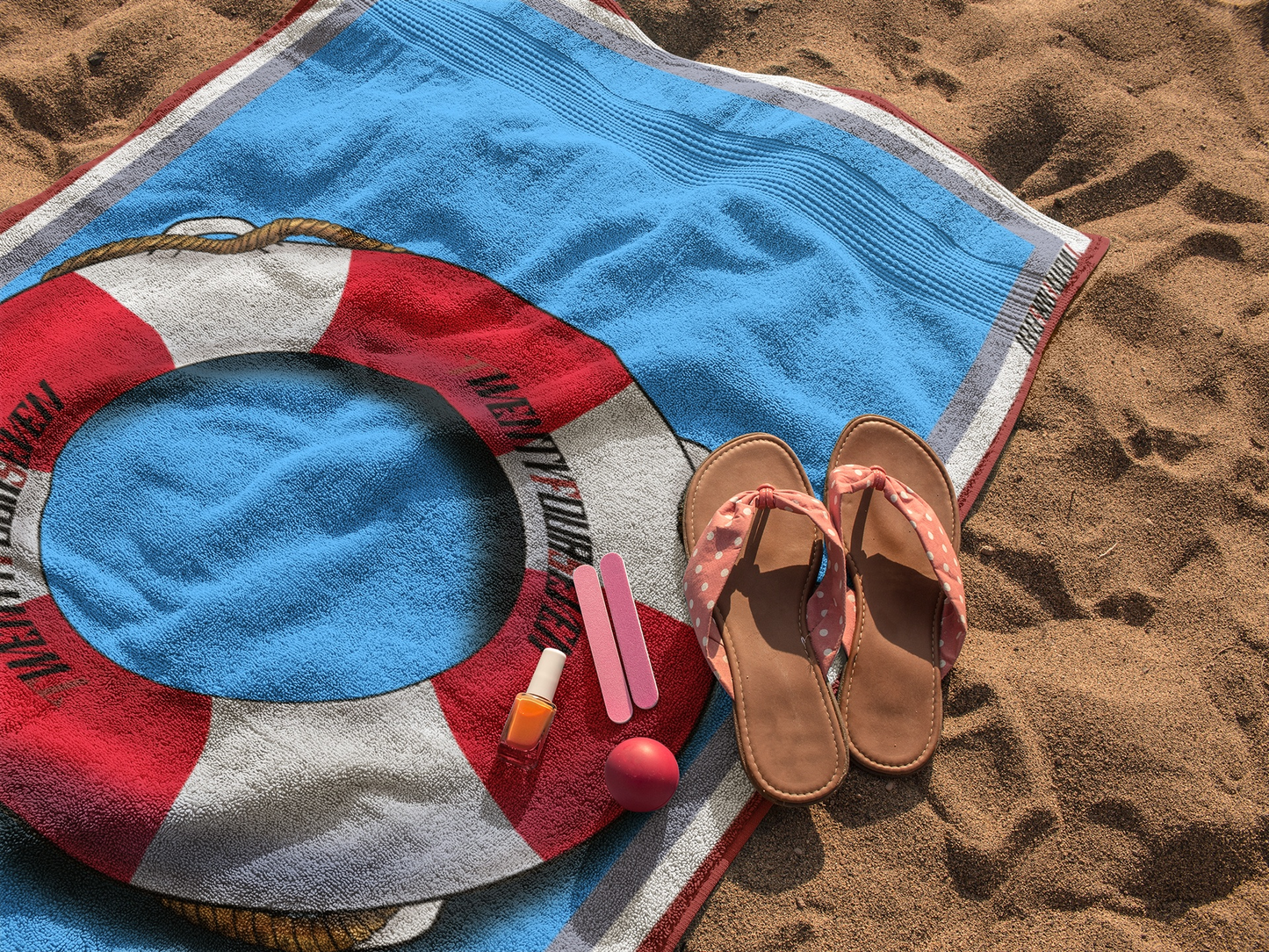 Nautical beach towel with a life belt design laid on the sand with summer sandals and accessories.