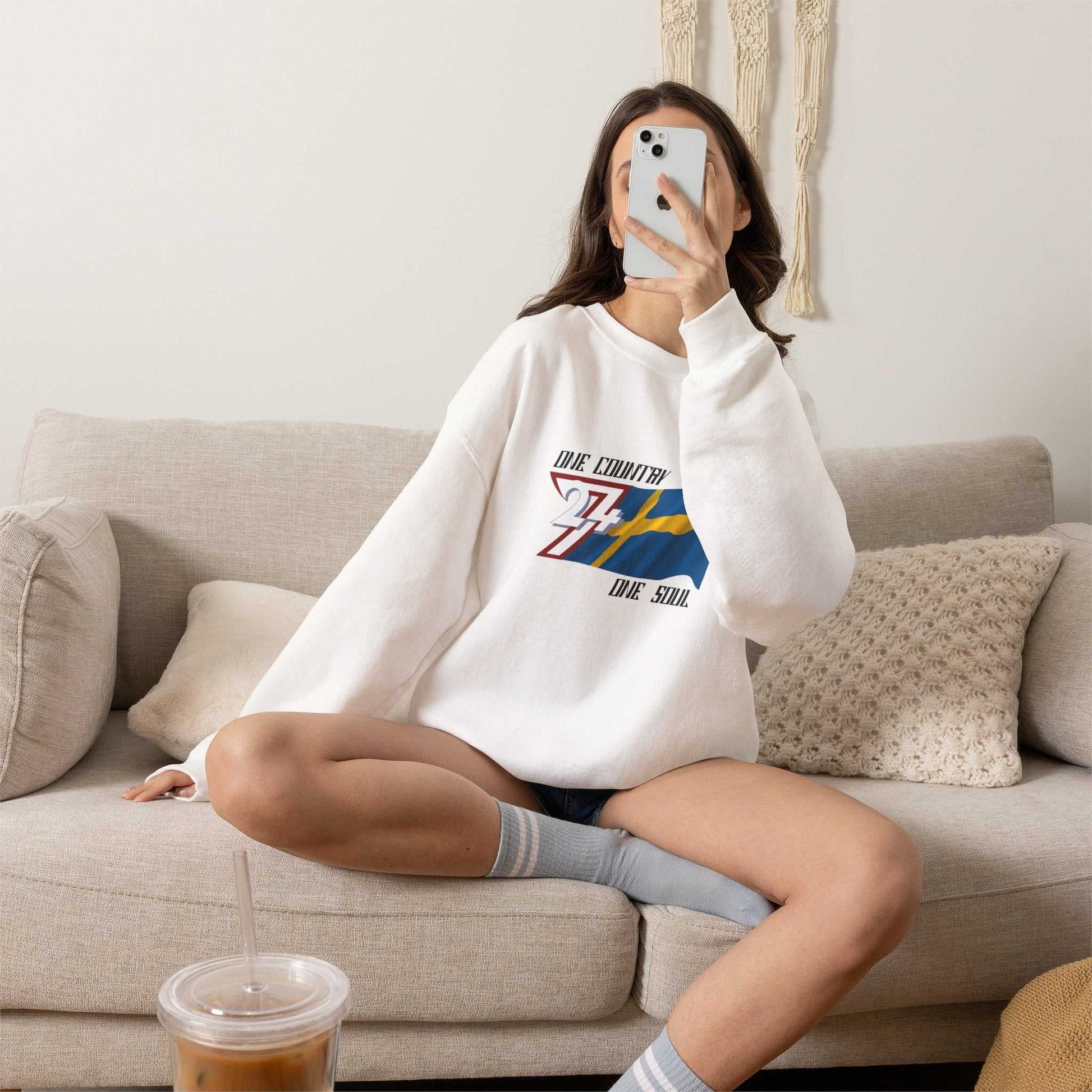 Female model wearing the white Sweden flag pullover, taking a selfie comfortably in the living room
