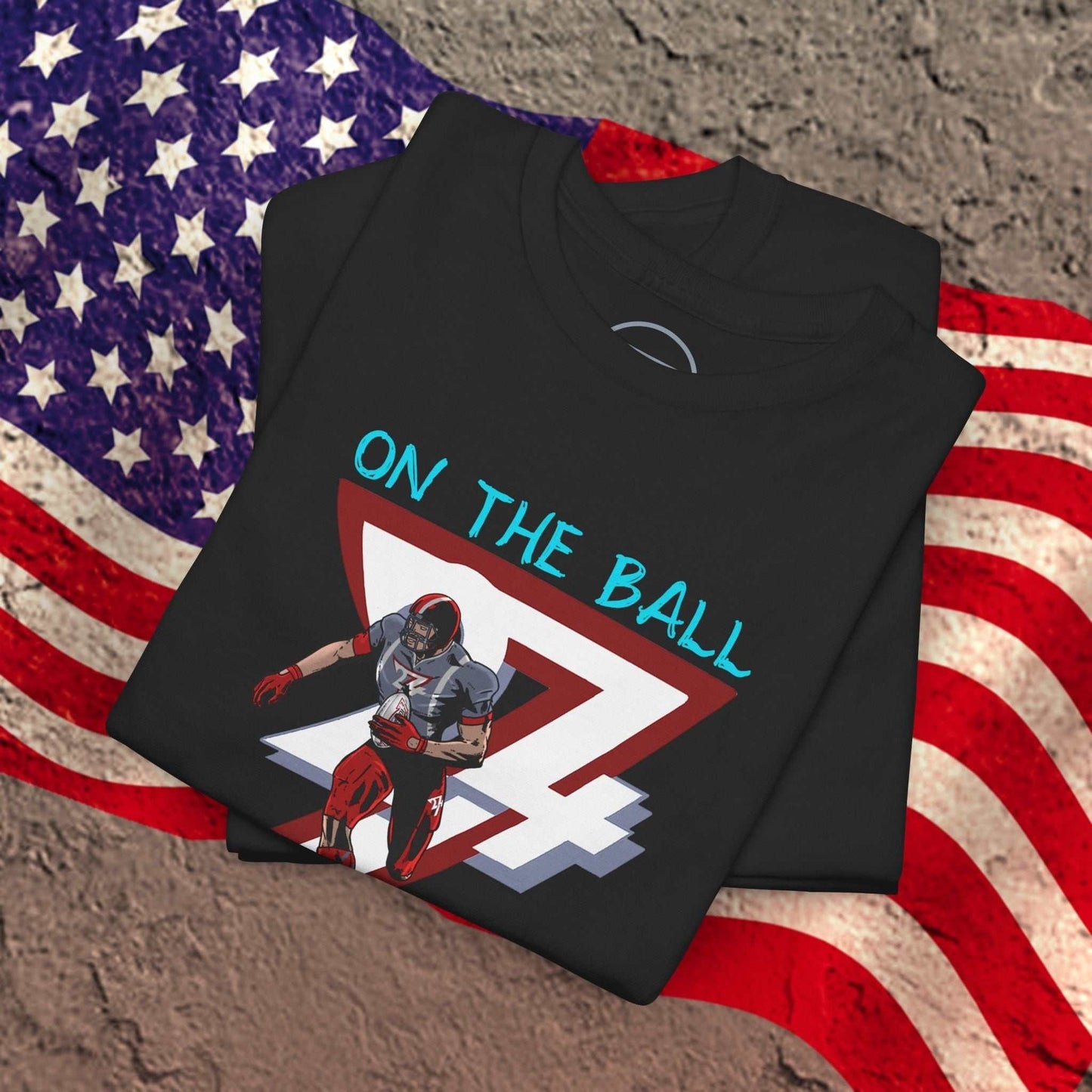 Black American football tee with hand-drawn comic-style graphic of a player and "On The Ball" text, laid on a textured surface with an American flag background.