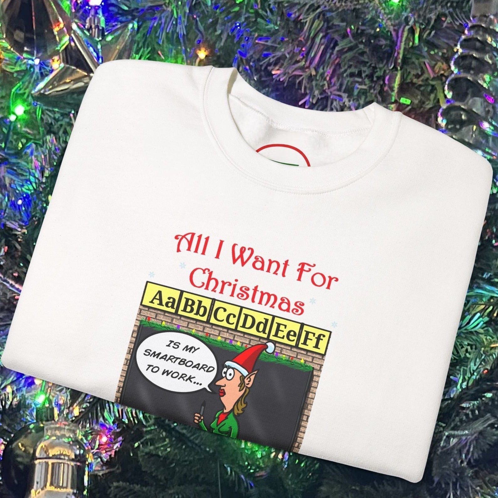 White teacher Christmas sweatshirt with funny cartoon comic design and holiday classroom theme folded on decorated Christmas tree background