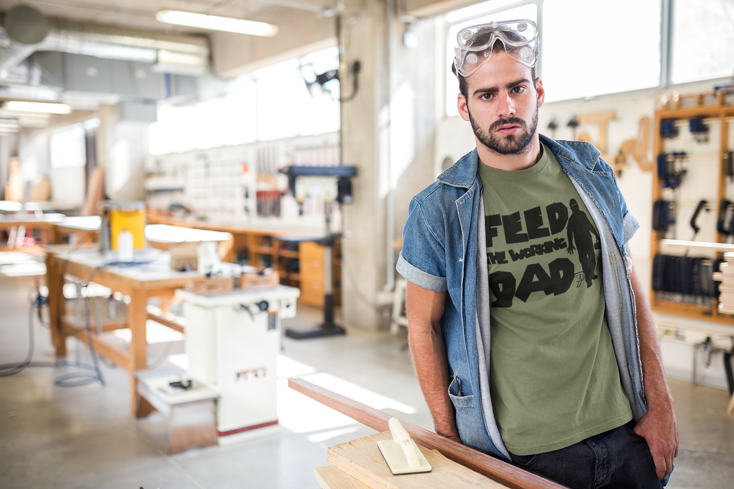 Male model styling the army green funny tradesman zombie dad tee with the black parody print, standing in a bright carpentry and woodworking shop.