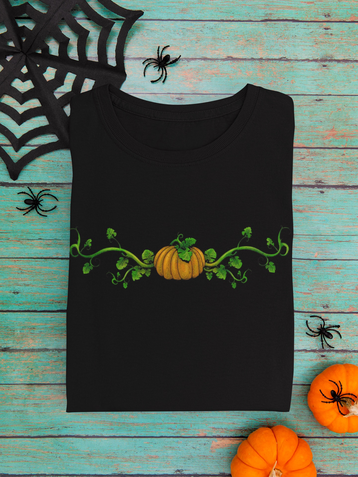 Black t-shirt with a pumpkin and vine design on a wooden surface with Halloween decorations.
