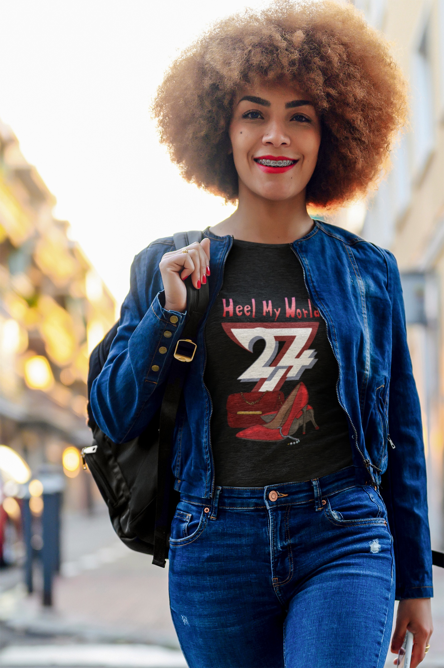 Female model wearing the black elegant high heel graphic tee under a denim jacket, smiling while walking down a sunny city street with a backpack.