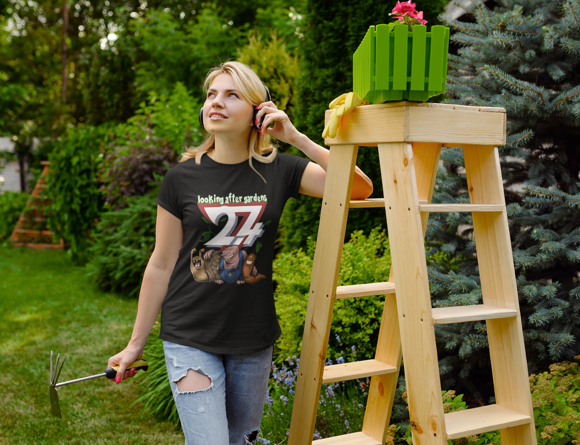 Female model wearing the black funny gardening shirt, listening to headphones while standing next to a wooden ladder in a lush backyard garden.