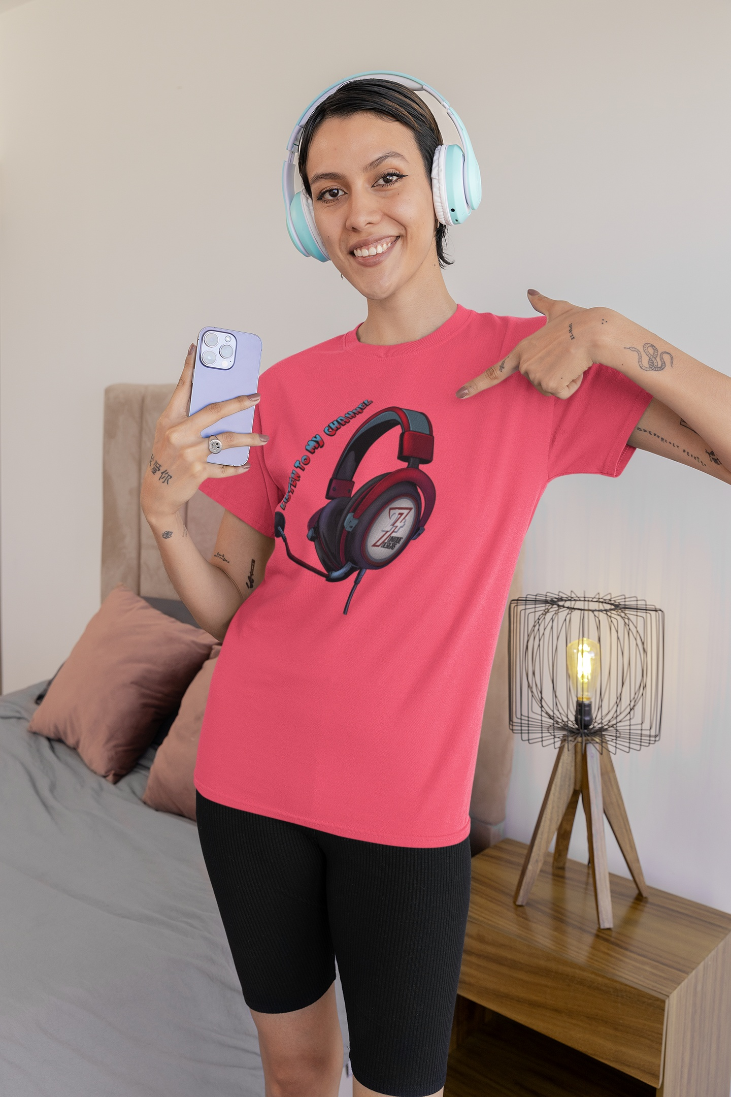 Female model styling the soft coral influencer graphic tee with the 'Listen To My Channel' art, pointing to the design while holding a phone in a relaxed bedroom setting.