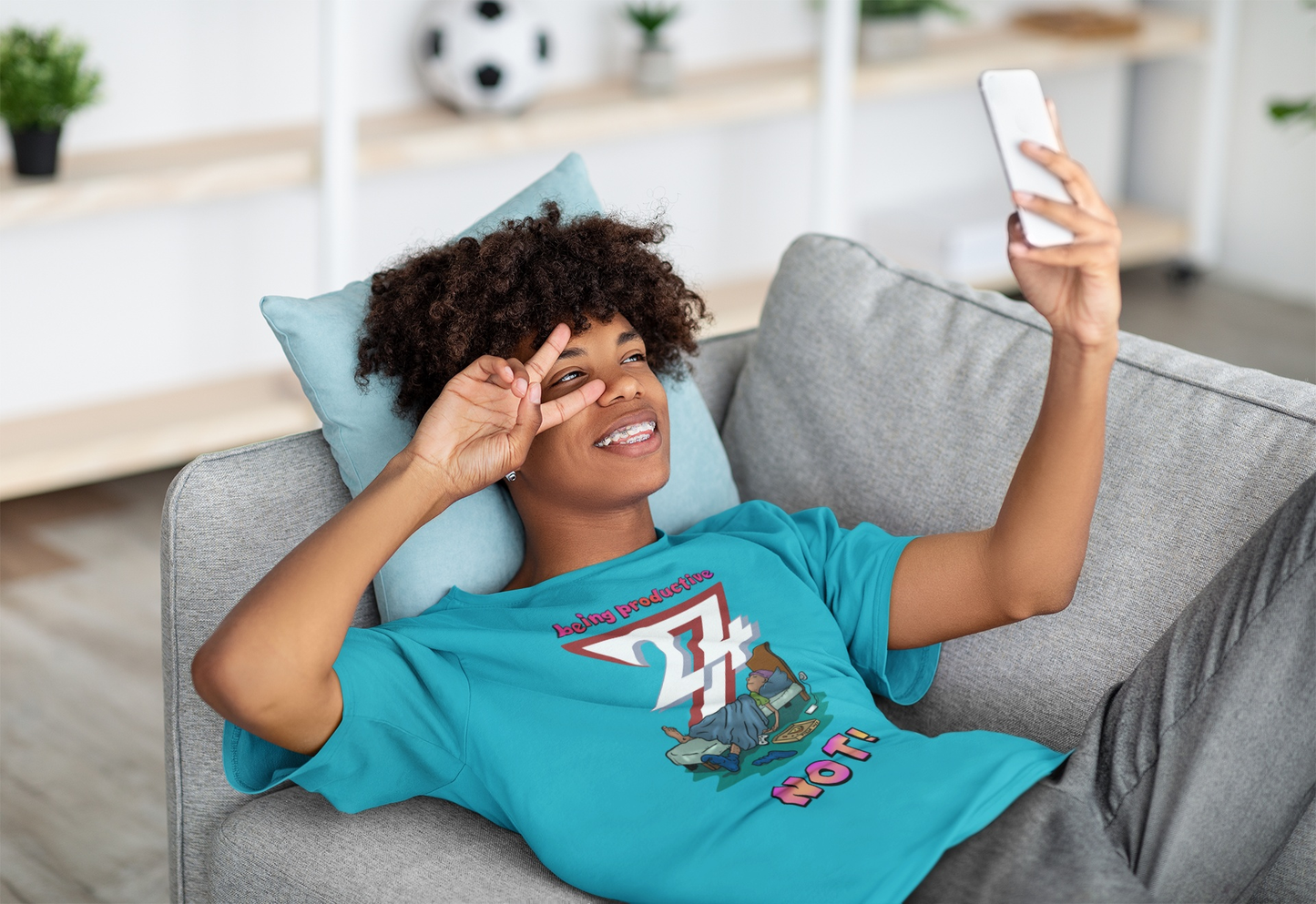 Young female model styling the teal anti-productivity graphic tee while relaxing on a sofa, showcasing the relaxed oversized fit, laying on her back and taking a selfie.
