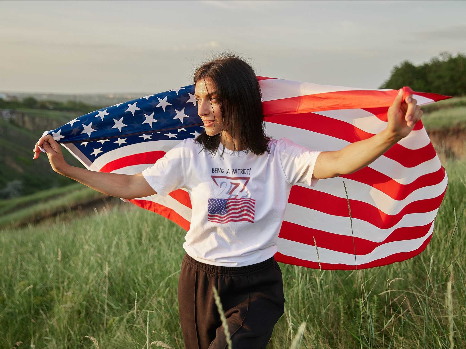 Female model showcasing the white American patriot flag t-shirt, letting a large US flag wave behind her in a lush, grassy outdoor field.