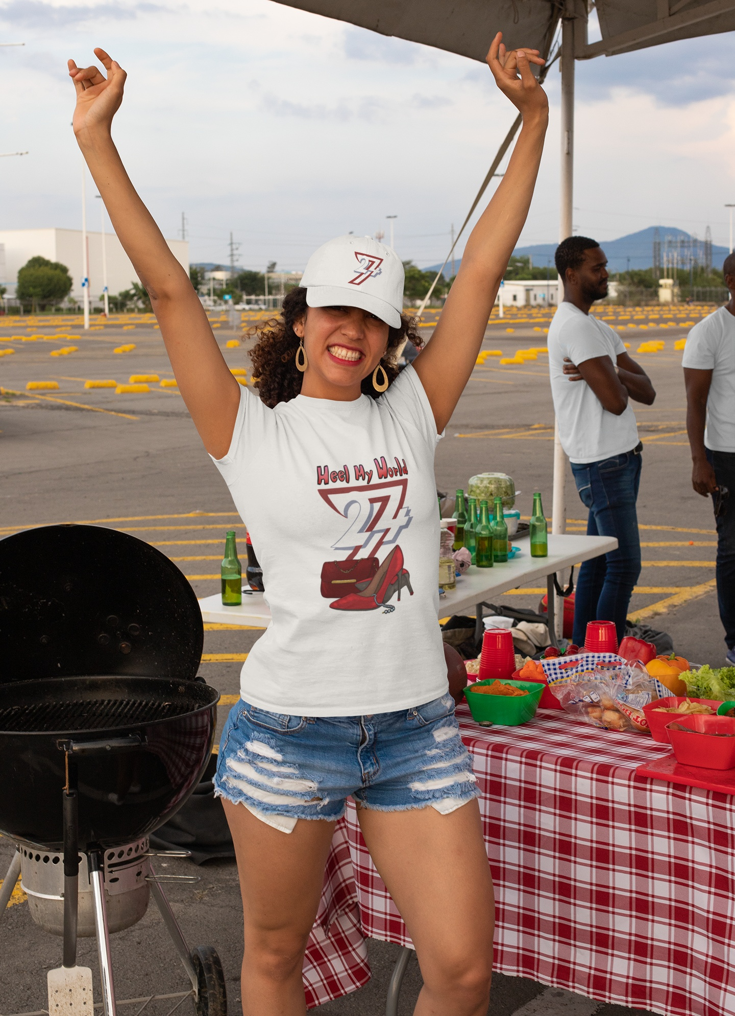 Cheerful female model wearing the white elegant high heel graphic tee and a 24/7 dad hat, raising her arms happily at an outdoor tailgate party.