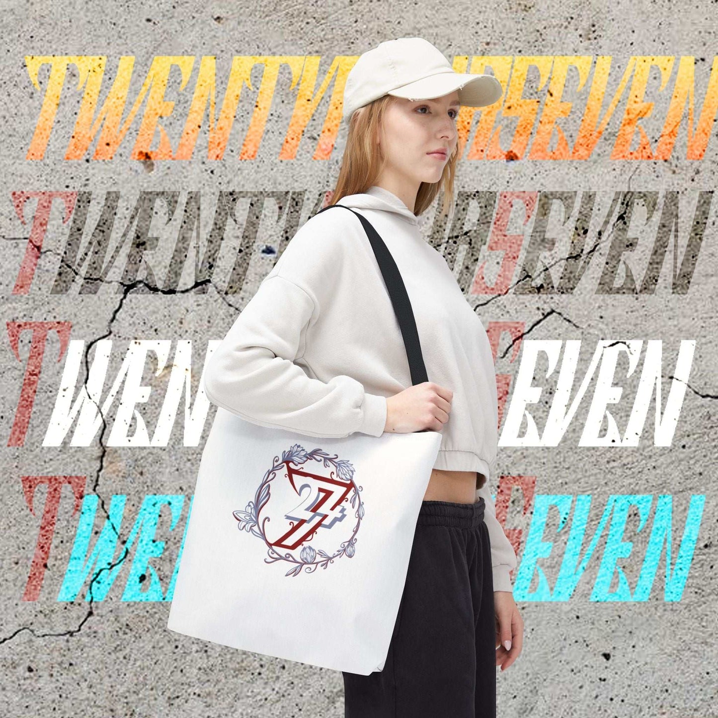 A woman wearing a white cap, white hoodie, and black sweatpants carries a white canvas floral tote bag with a black strap. The bag features a graphic of a red and blue 24/7 logo inside a floral wreath.