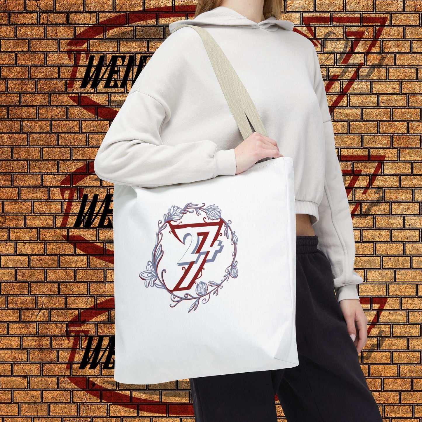 A person wearing a white cropped hoodie and black sweatpants carries a white canvas floral tote bag with a beige shoulder strap. The bag features a graphic print of a red and blue 24/7 unique designs logo inside a circular floral wreath.