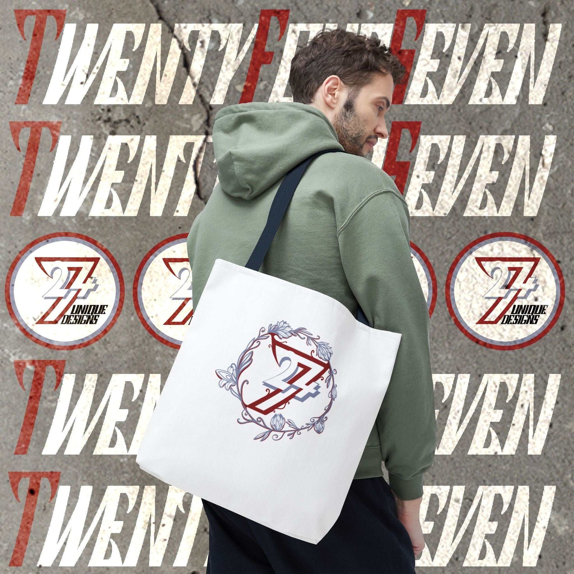 A man wearing an olive green hoodie carries a white canvas tote bag over his shoulder. The floral tote bag features a circular graphic with a stylized red and blue 24/7 monogram and floral detailing.