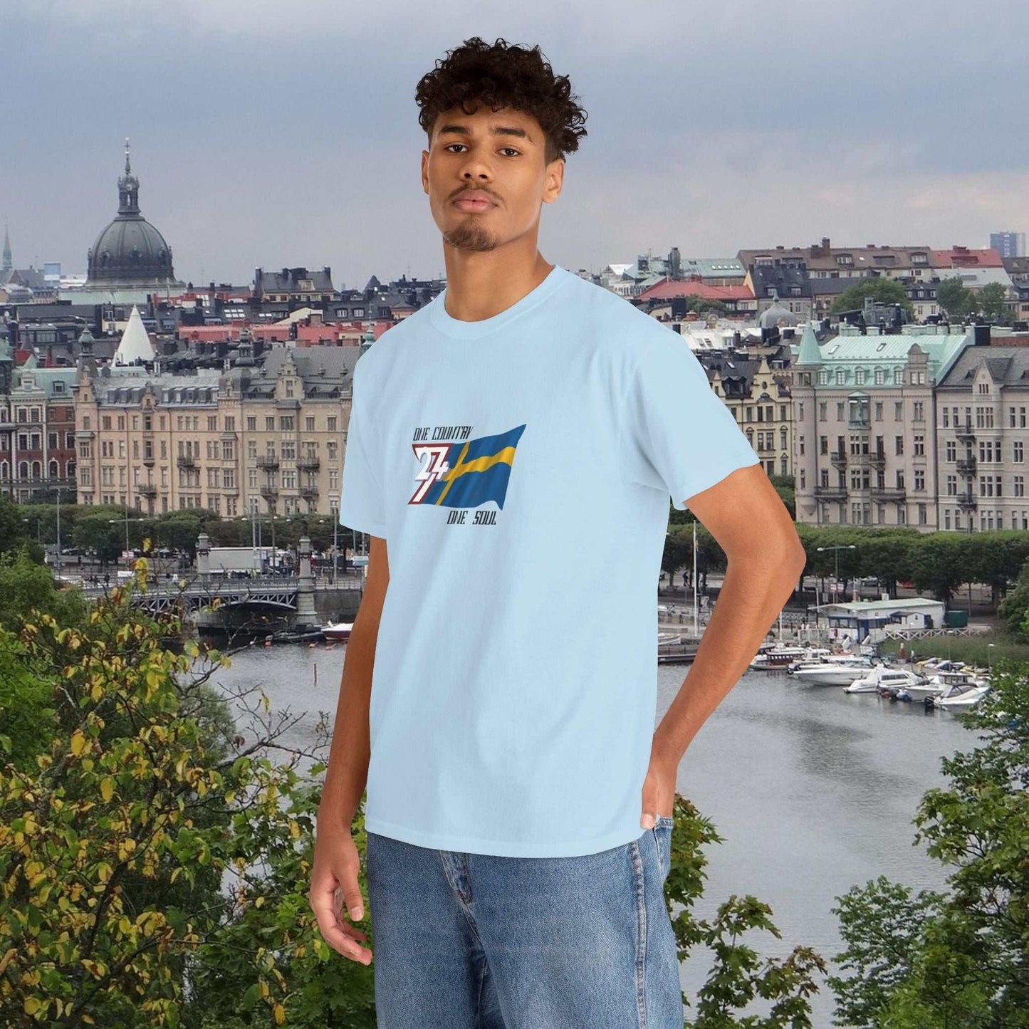 man wearing light blue Sweden flag graphic tee with hand-drawn Swedish flag and text standing outdoors with cityscape background