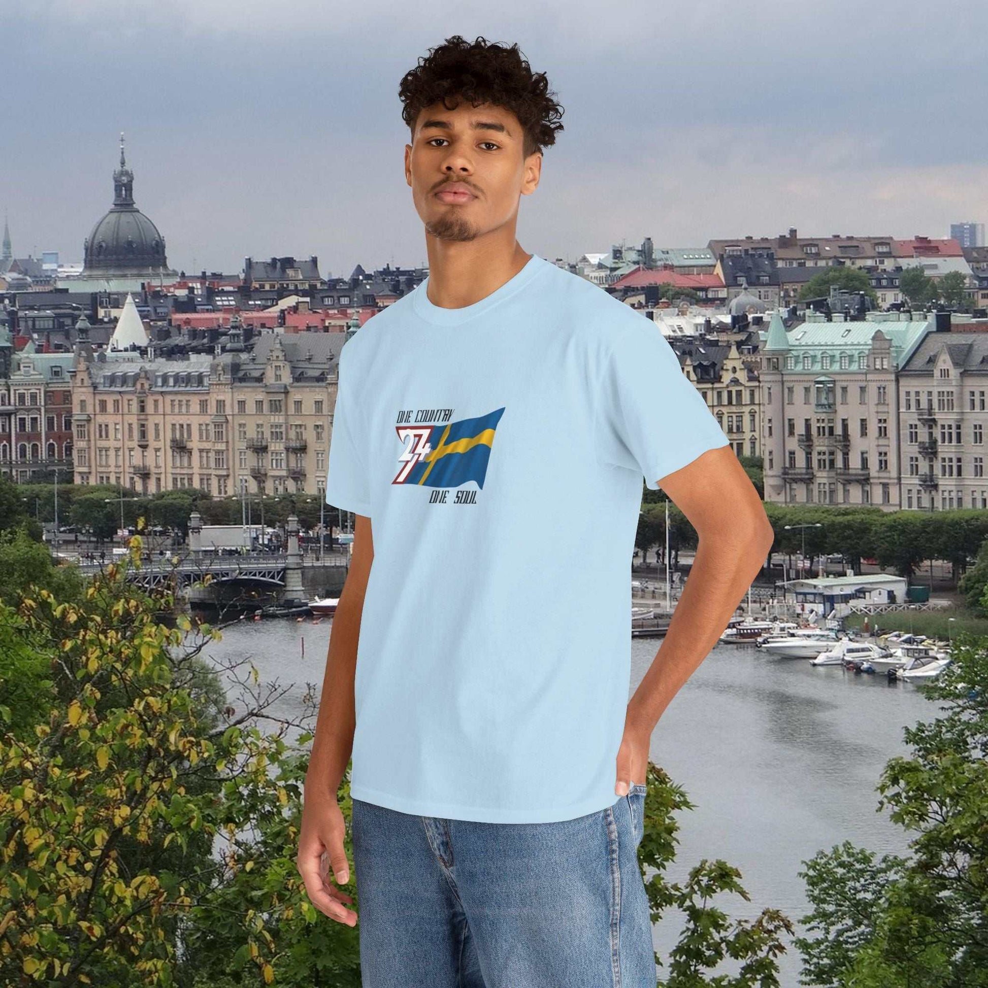 man wearing light blue Sweden flag graphic tee with hand-drawn Swedish flag and text standing outdoors with cityscape background