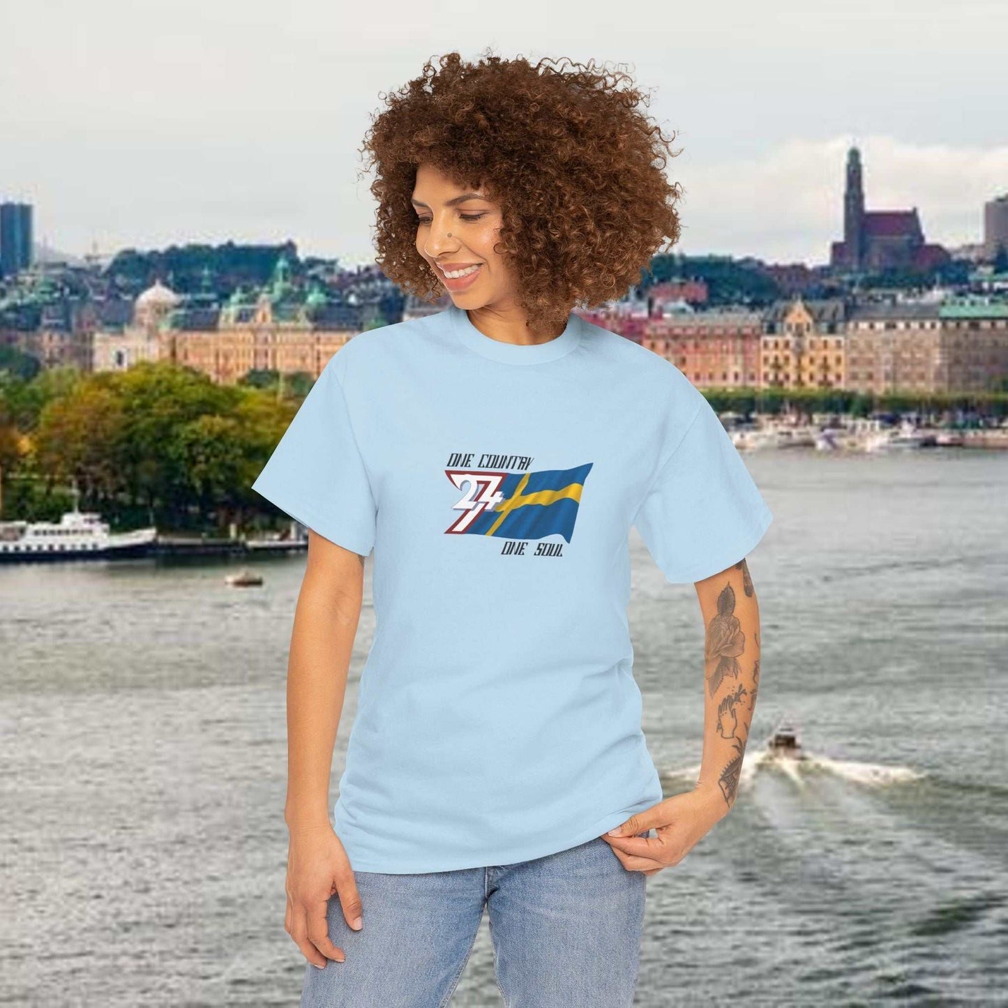 Woman wearing light blue Sweden Flag Graphic Tee with hand-drawn Swedish flag and text, standing near waterfront cityscape.