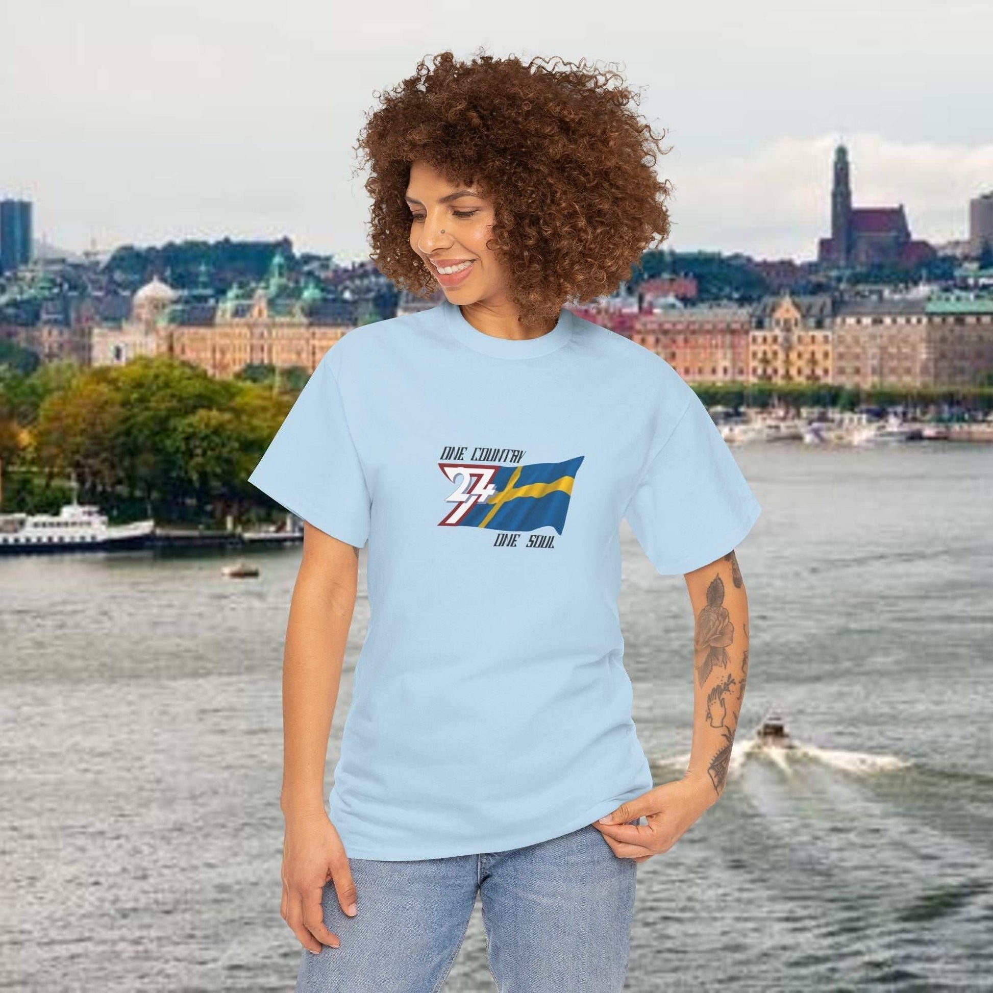 Woman wearing light blue Sweden Flag Graphic Tee with hand-drawn Swedish flag and text, standing near waterfront cityscape.