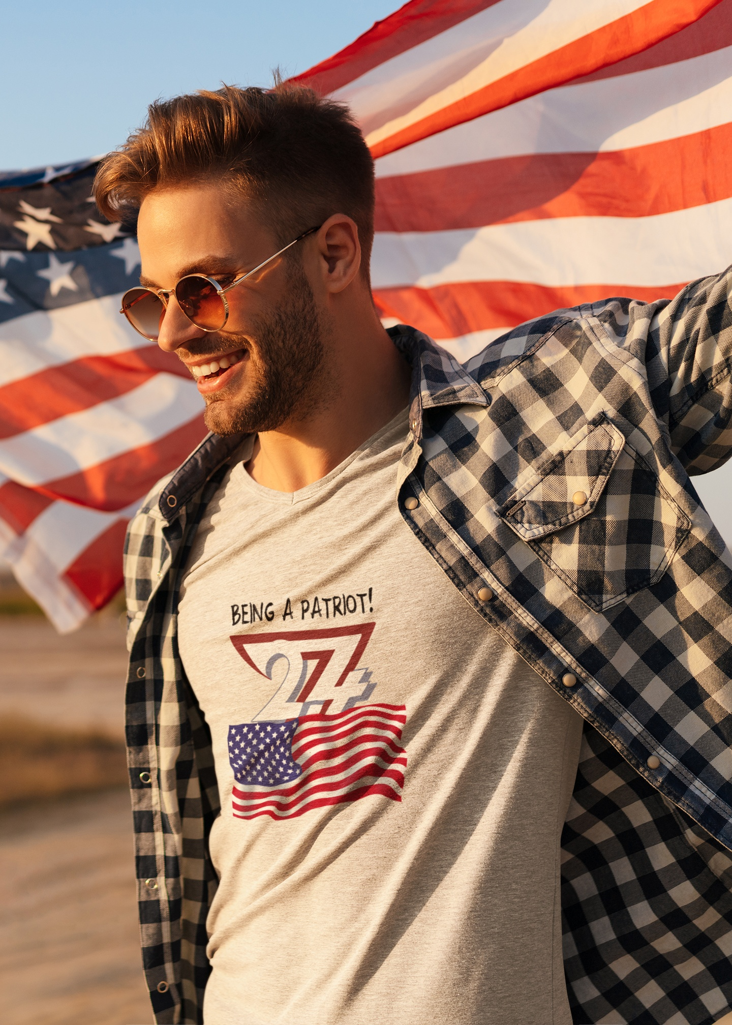 Smiling male model wearing the beige American patriot flag t-shirt under a flannel shirt, proudly holding a large American flag outdoors at sunset.