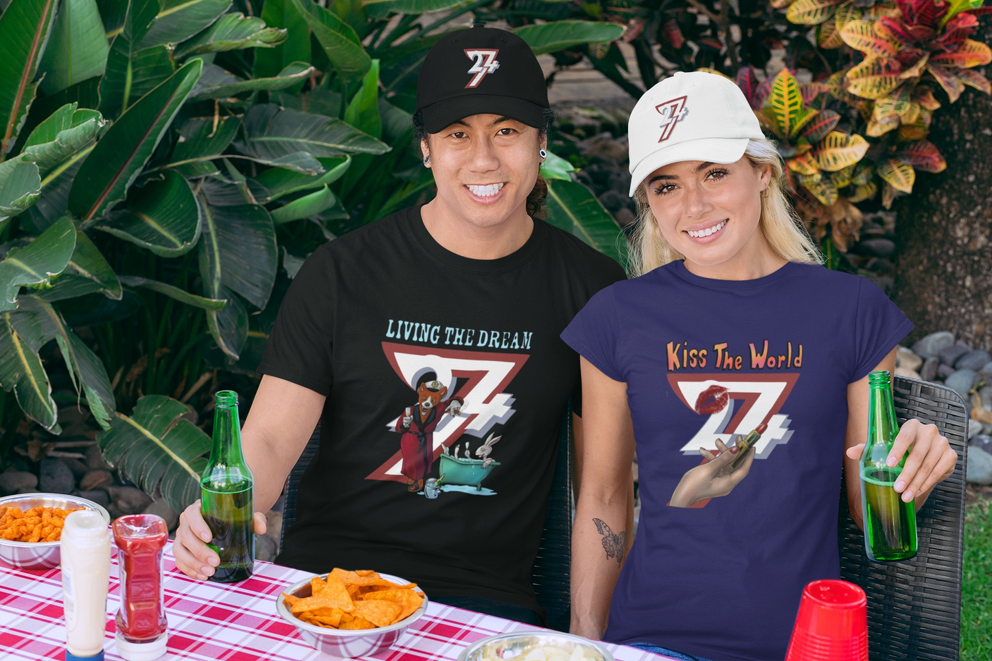 Male model wearing the black funny bachelor party shirt featuring the Living The Dream fox graphic, sitting at an outdoor BBQ alongside a female model styling the 24/7 Kiss The World tee and dad hats.