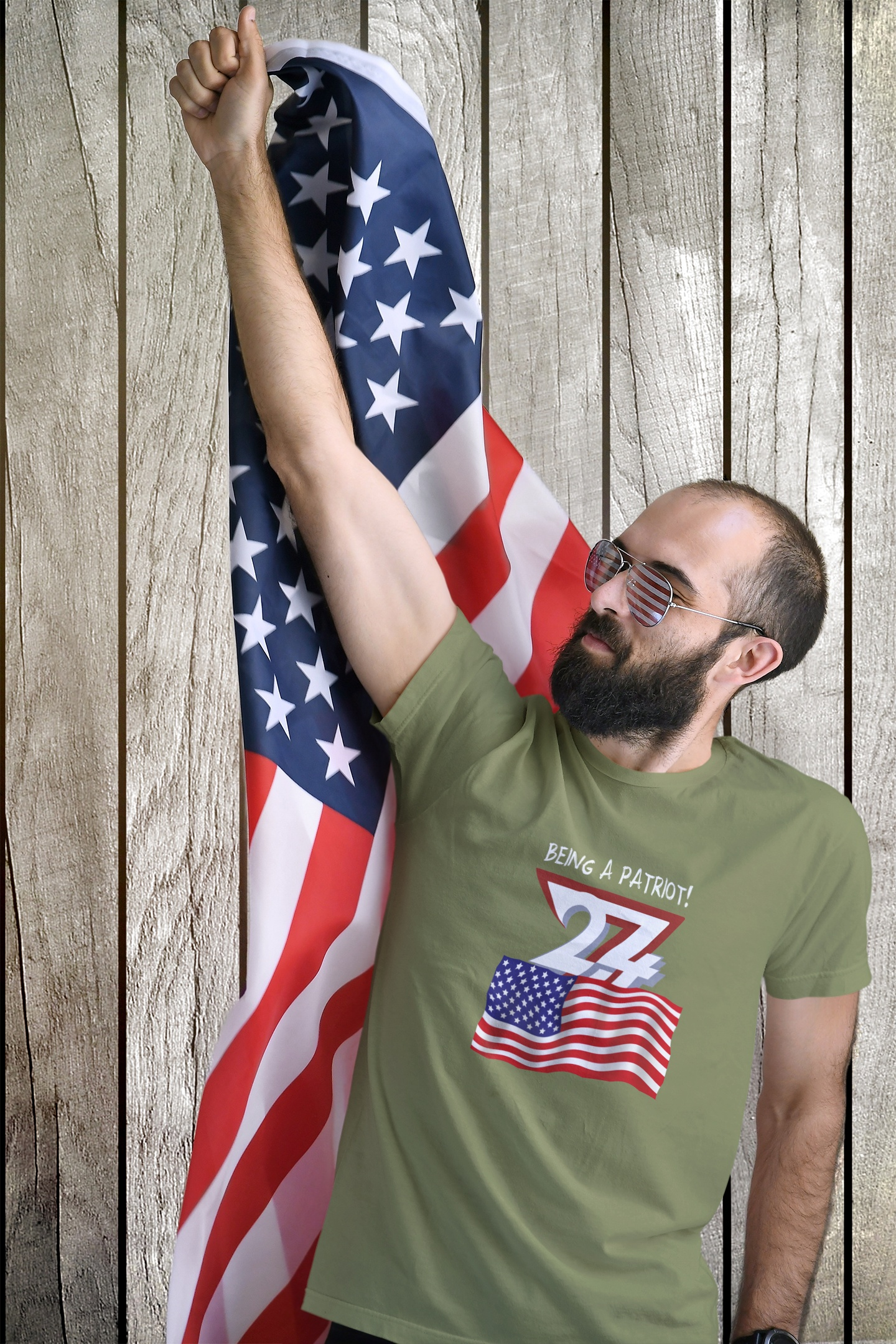 Male model wearing the olive green Independence Day clothing, raising a draped American flag with one hand against a rustic wooden backdrop.