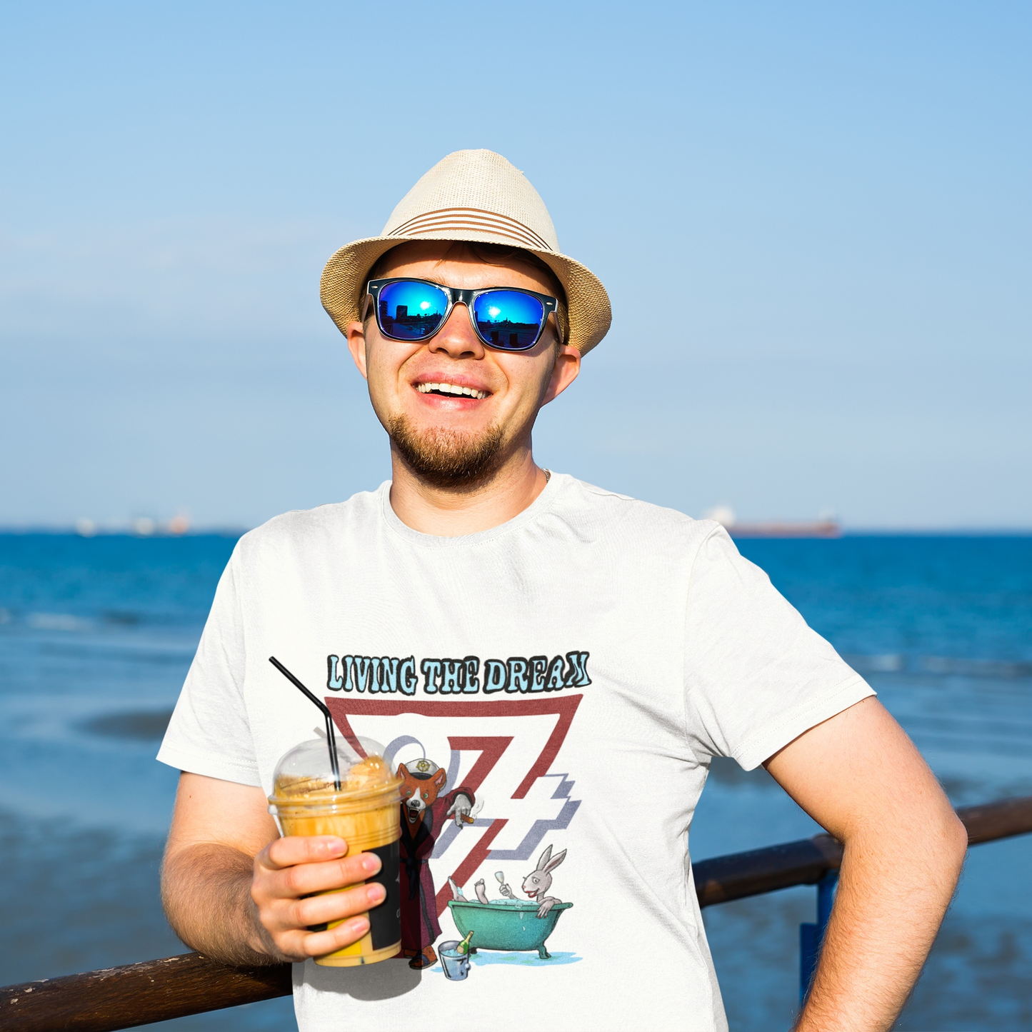 Male model wearing the white Living The Dream fox tee showcasing the colorful animal cartoon graphic, smiling with a coffee on a bright, sunny ocean pier.
