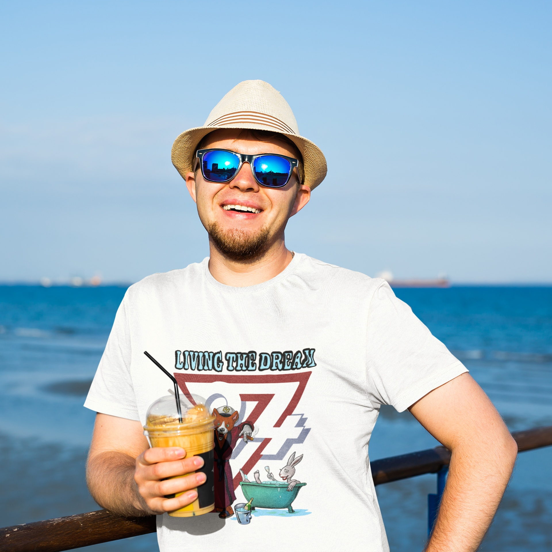 Male model wearing the white Living The Dream fox tee showcasing the colorful animal cartoon graphic, smiling with a coffee on a bright, sunny ocean pier.