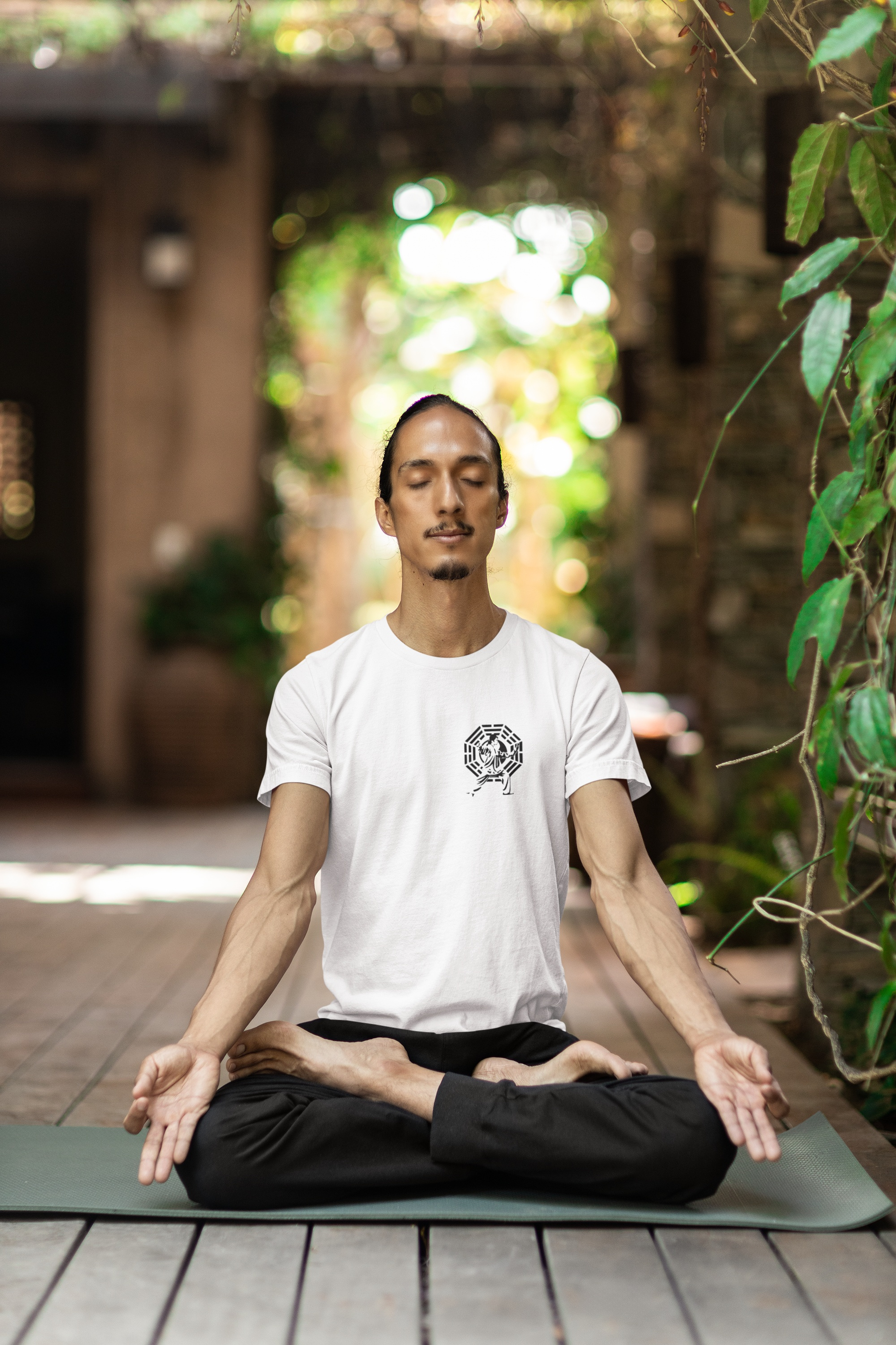 Man wearing the white Tai Chi martiak arts tee meditating on a mat outdoors with greenery in the background