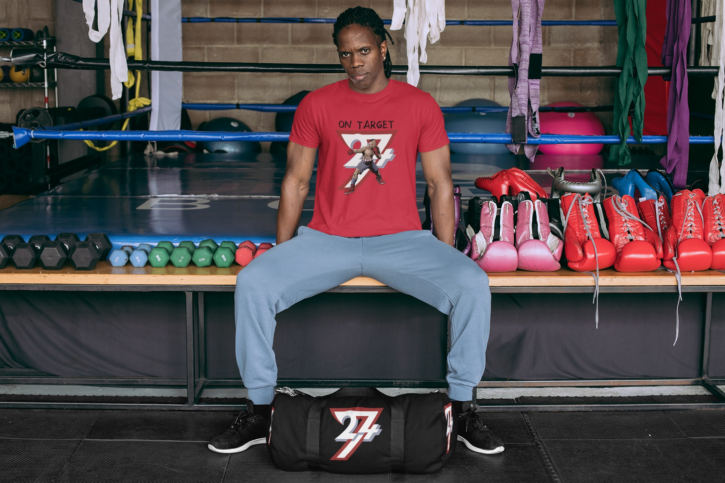 Tough-looking male athlete sitting on a bench at a boxing gym with his 24/7 duffle bag, wearing the red vintage boxing graphic tee surrounded by boxing gloves and weights.