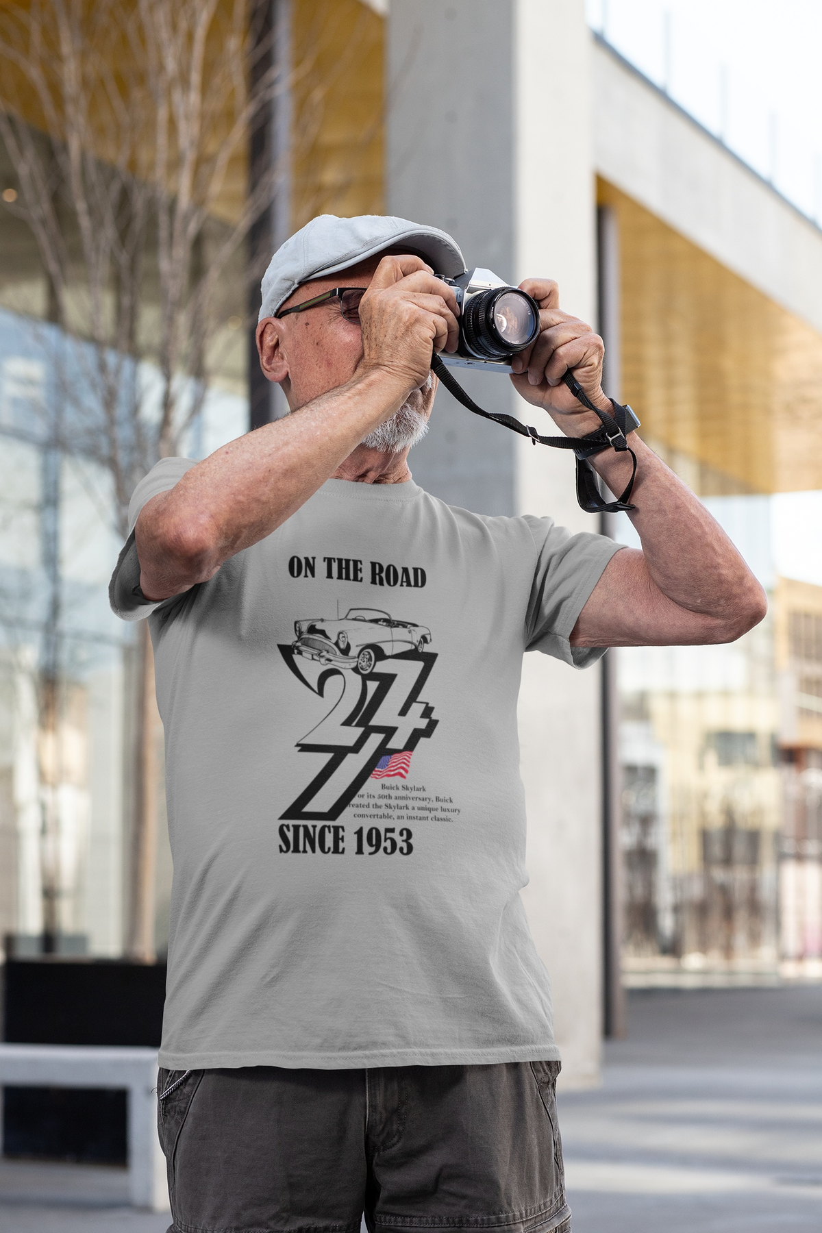 Older man wearing a cap and holding a vintage camera to his eye, modeling the sport gray vintage American car shirt outdoors.