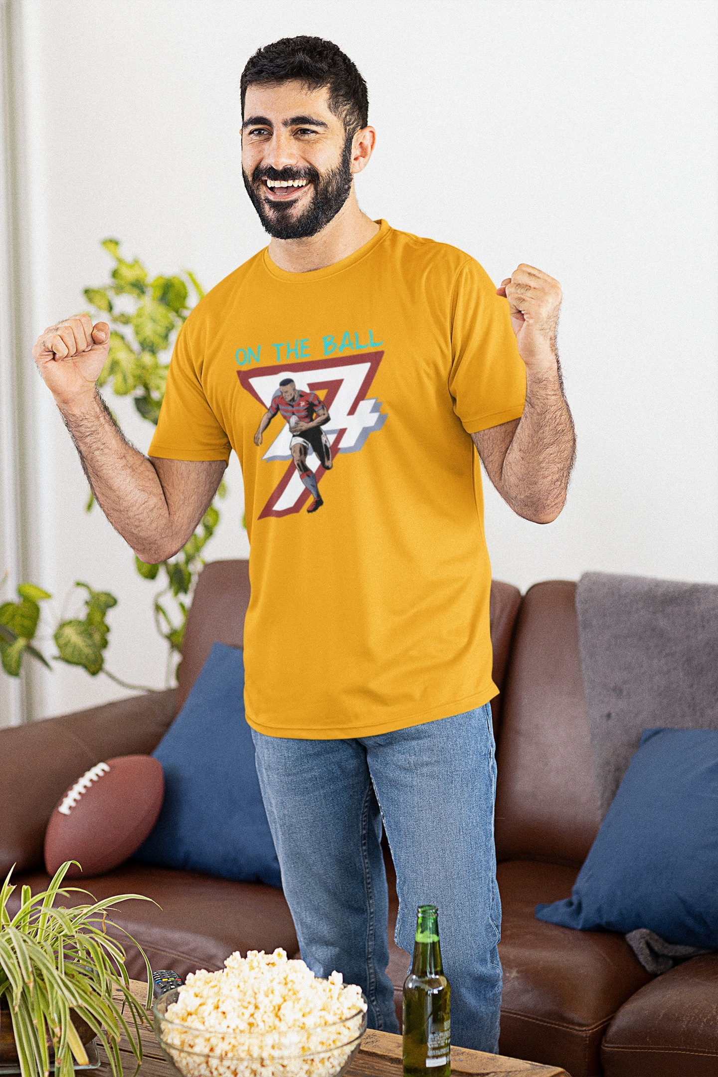 Excited sports fan wearing the gold comic-style rugby graphic tee, cheering and watching the big game at home with snacks and drinks.