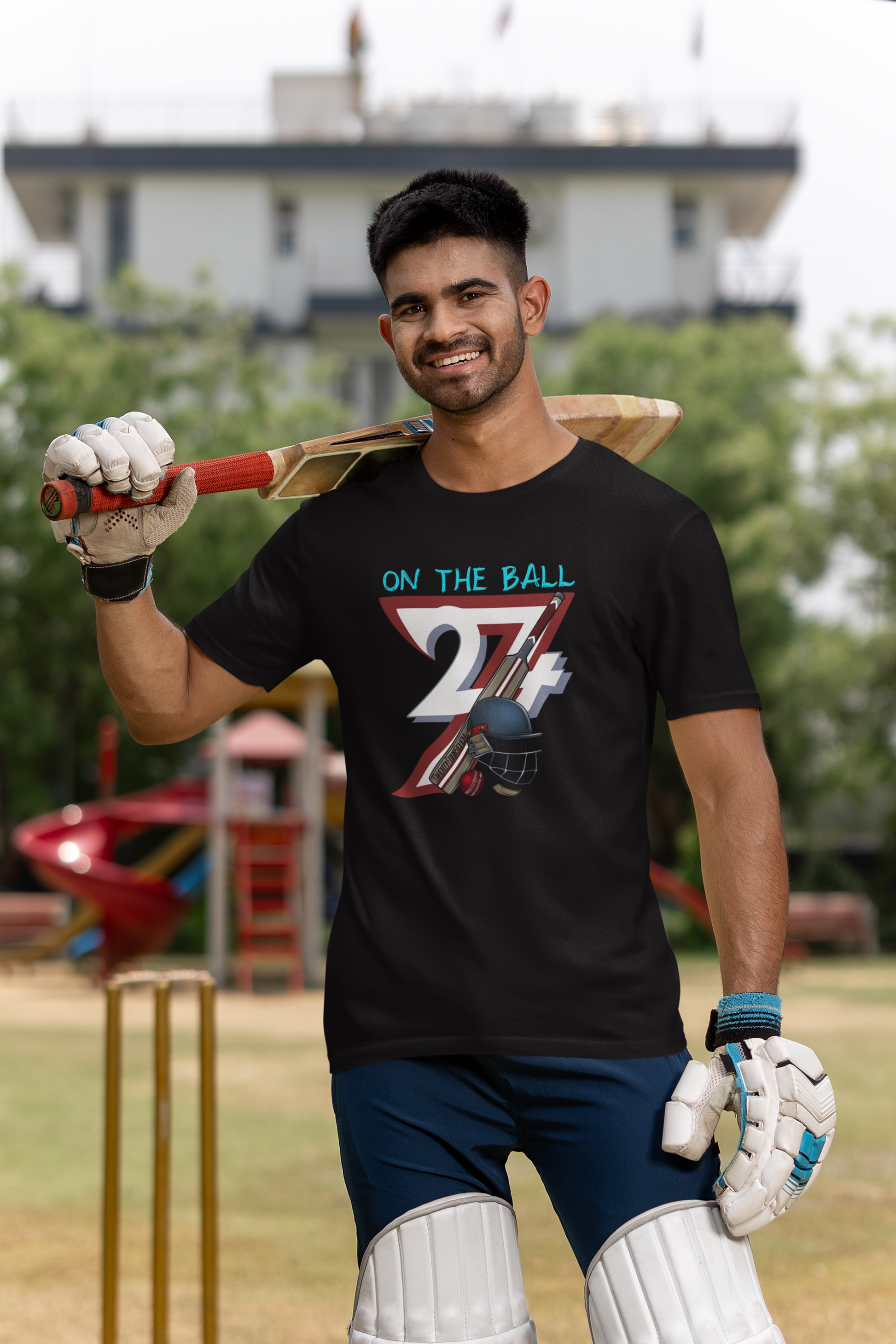 Smiling male cricket player standing on a field wearing batting pads and gloves, casually resting a bat on his shoulder while wearing the black retro cricket shirt.