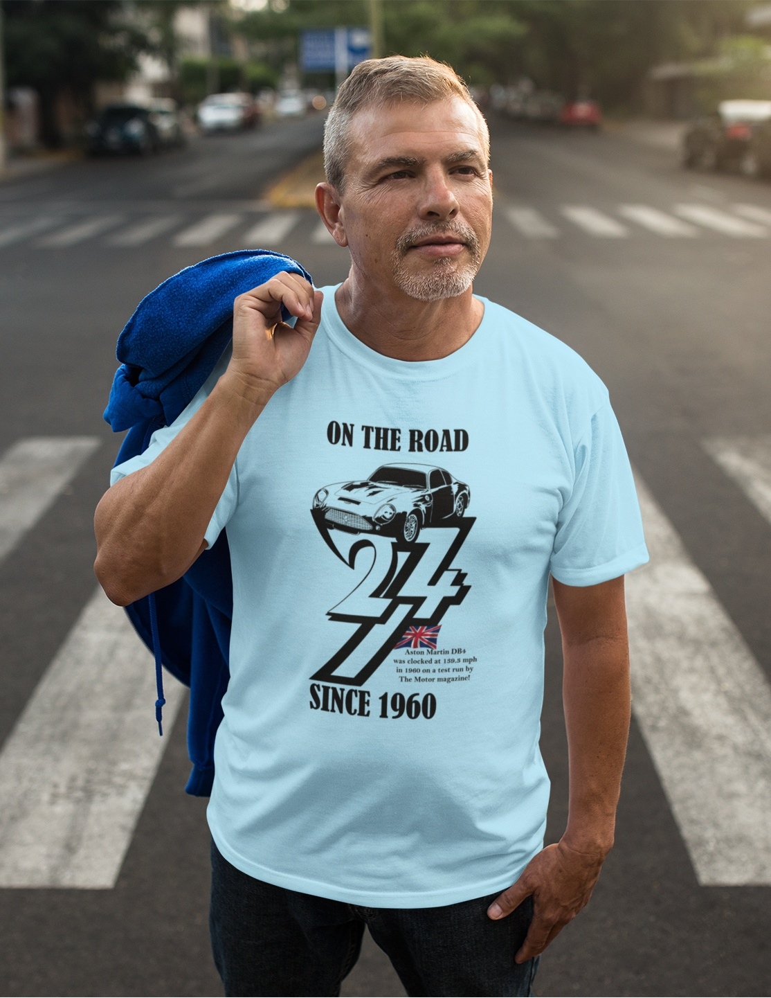 Handsome older man standing confidently in a city crosswalk with a blue jacket slung over his shoulder, modeling the light blue 1960 classic car birthday tee.