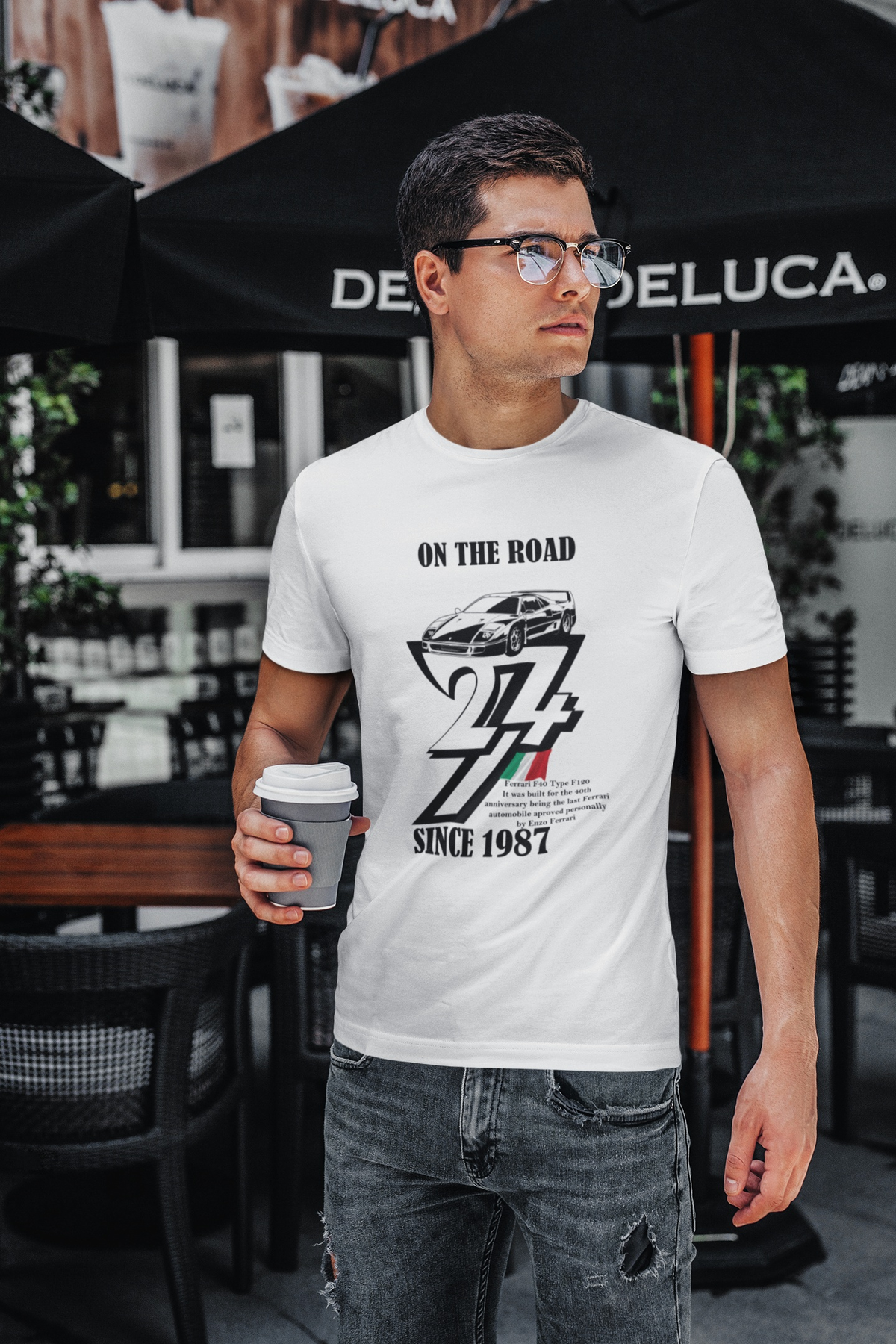 Stylish young man wearing glasses and distressed jeans standing outside a cafe holding a coffee cup, modeling the crisp white 1987 Ferrari F40 graphic tee.