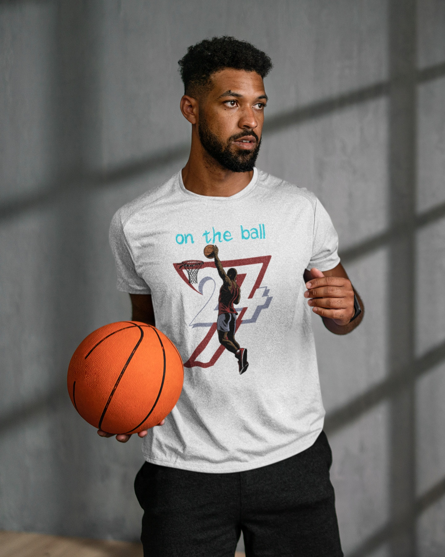 Male athlete standing casually holding a basketball in a modern space with dramatic window shadows, wearing the sport gray hand-drawn streetwear shirt to showcase its relaxed, everyday fit.