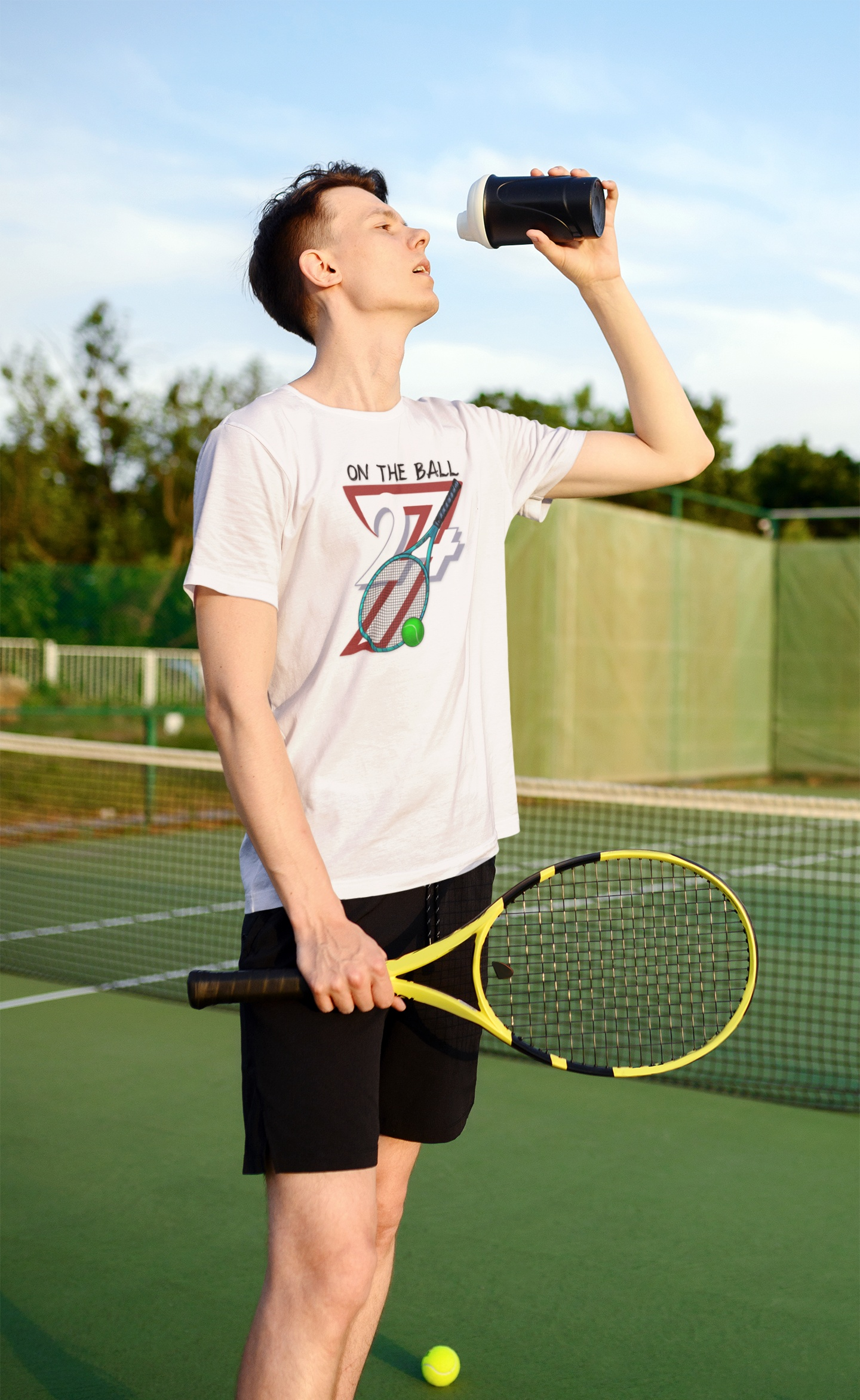 Young male tennis player taking a water break on an outdoor green hardcourt, holding his racket and wearing the natural 90s retro sports t-shirt.