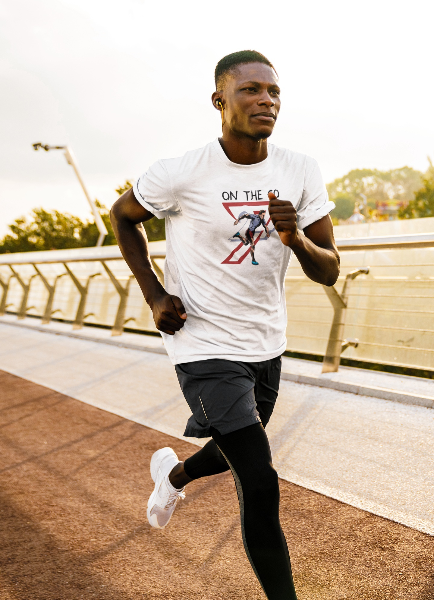 Focused young male athlete wearing the white comic style running shirt and black athletic leggings, jogging across a modern sunlit bridge while listening to music.