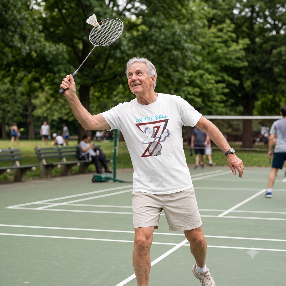 Smiling older gentleman enjoying a casual game of badminton in a leafy public park, preparing to return a shuttlecock while wearing the crisp white digital art sports t-shirt.