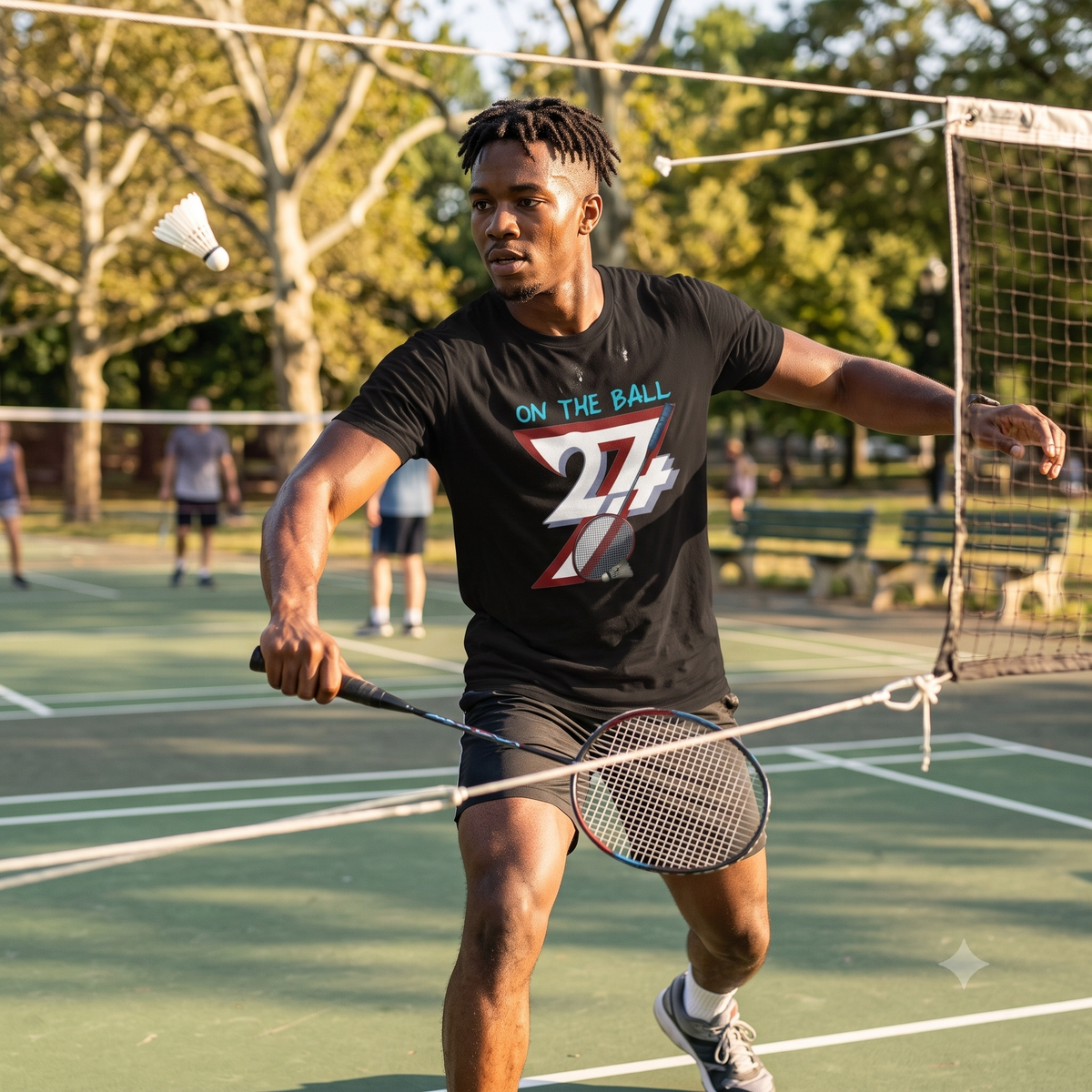 Athletic young man in an intense badminton rally on a sunny outdoor park court, wearing the black comic-style badminton graphic tee as he prepares to hit a mid-air shuttlecock.