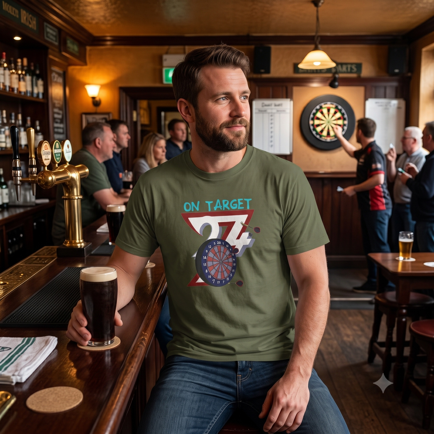 Handsome man sitting at a warmly lit pub bar with a pint of stout, wearing the army green dart league apparel tee while a game of darts is played in the background.