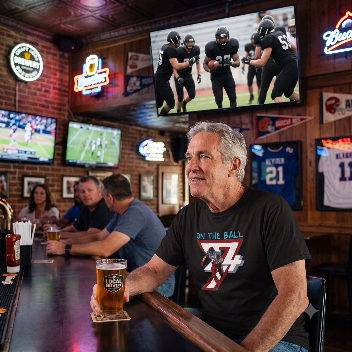 Smiling older man sitting at a lively sports bar holding a glass of local beer, wearing the black comic-style American football graphic tee while a football game plays on the TV behind him.