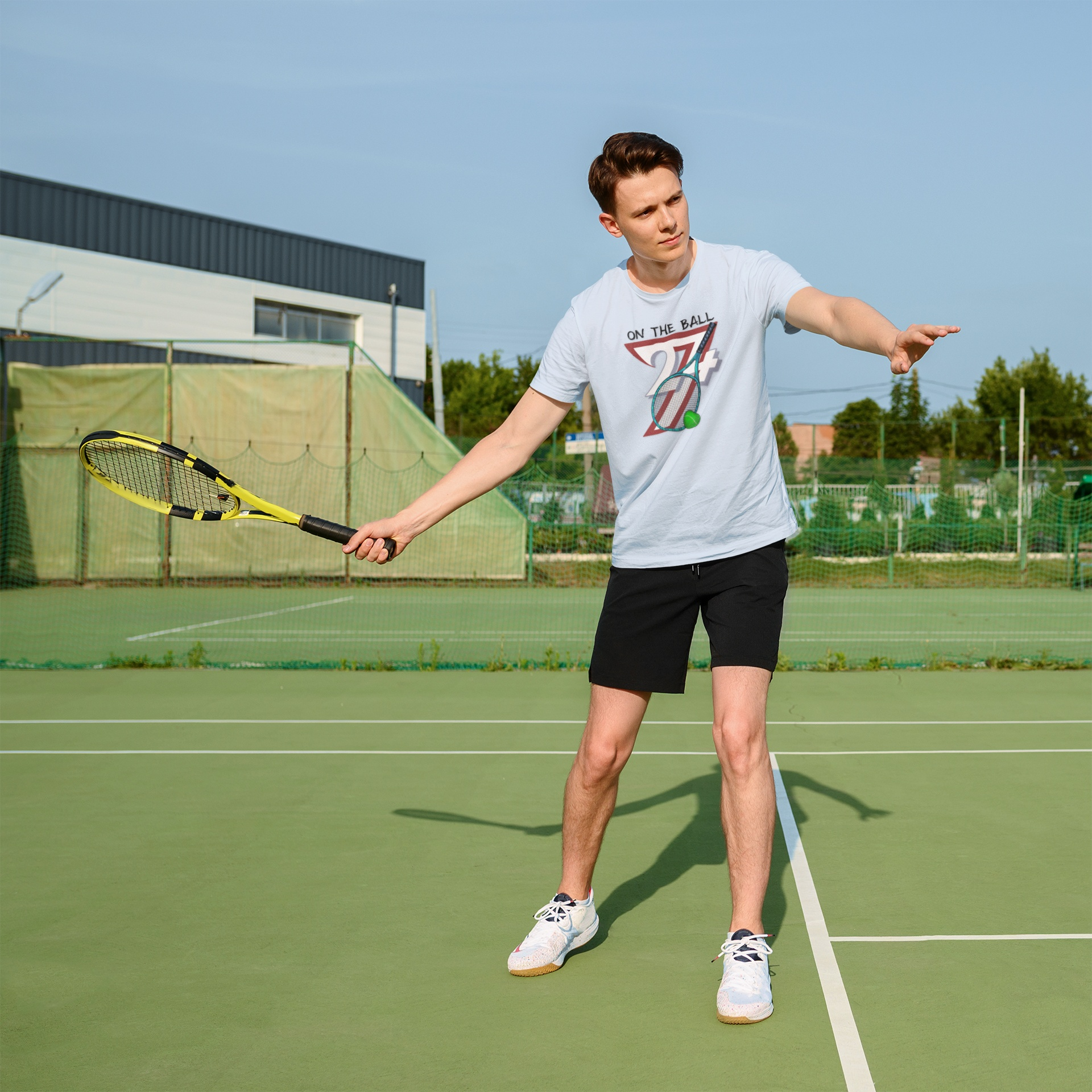 Young male athlete wearing the light blue vintage tennis graphic tee, following through on a powerful forehand swing during a sunny afternoon practice session on an outdoor hardcourt.