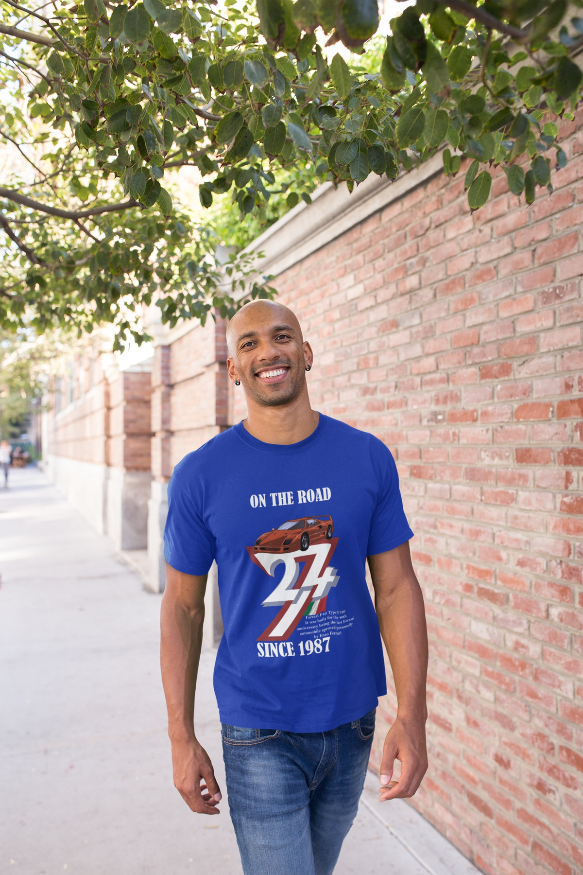 Happy bald man walking down a sunny brick-lined street under a tree, modeling the royal blue vintage Italian sports car shirt.