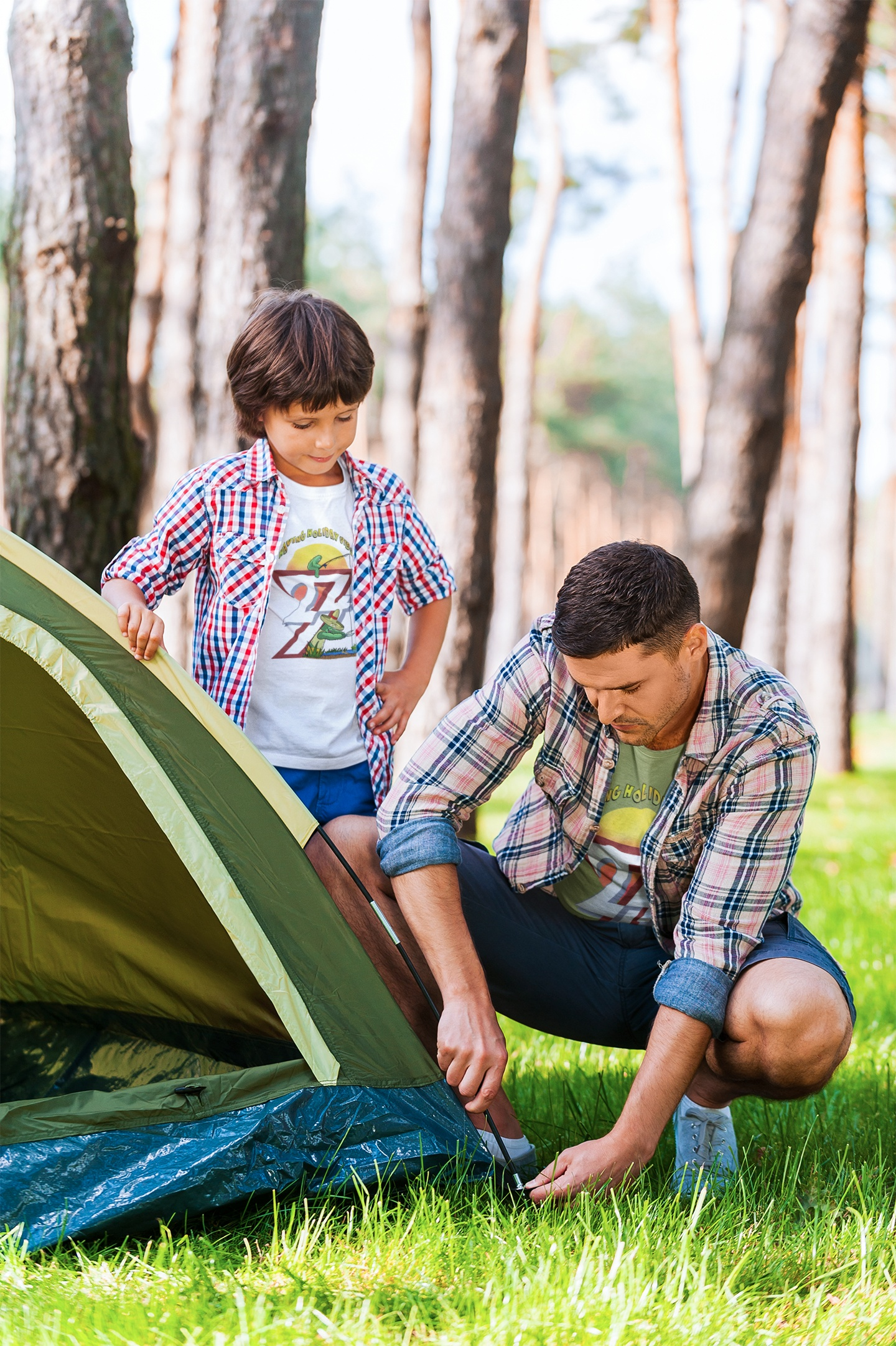 Father and son setting up a green camping tent. The father is wearing a lime green funny camping shirt under an open flannel, and the son is wearing a crisp white funny fishing tee.