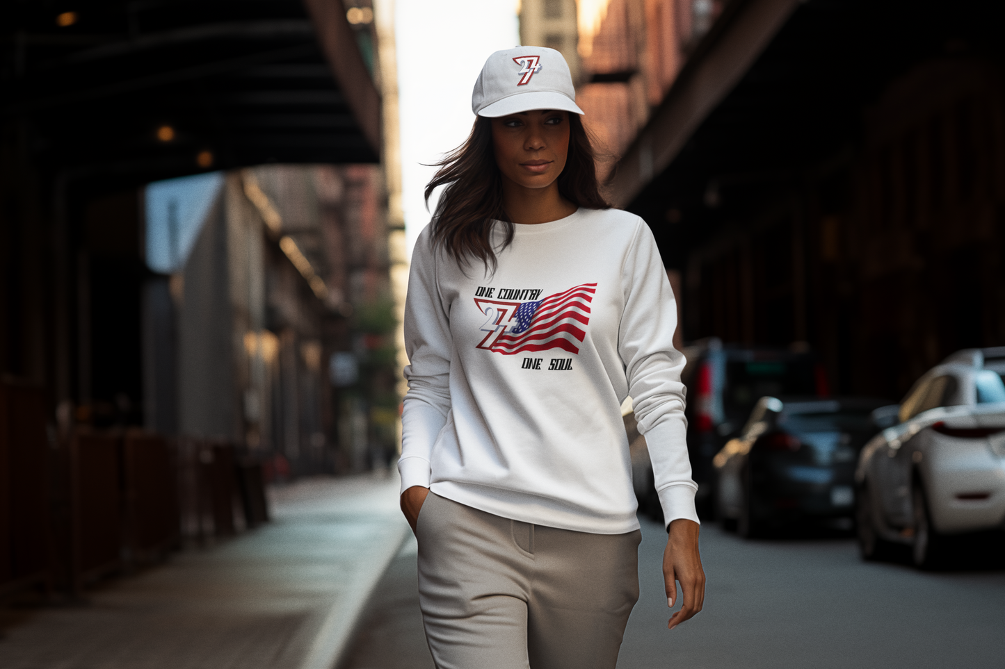 Female model wearing a white sweatshirt with an American flag design and a 24/7 Unique Designs white cap, walking on a street.