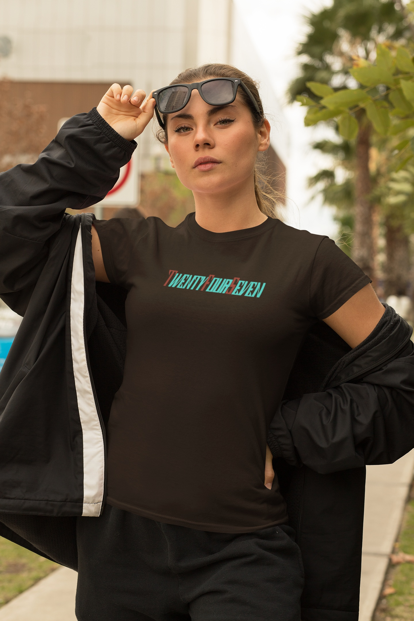 A female model wearing a dark chocolate premium streetwear t-shirt with the retro-futuristic logo, against an urban backdrop.