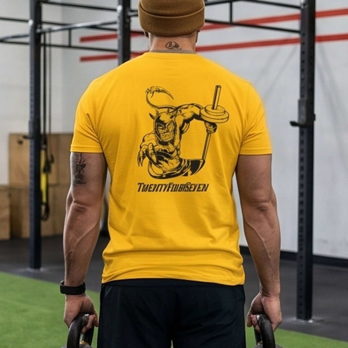Male model wearing a beanie and the gold hand-drawn fitness apparel tee, seen from the back holding kettlebells in a training facility, emphasizing the bold graphic.
