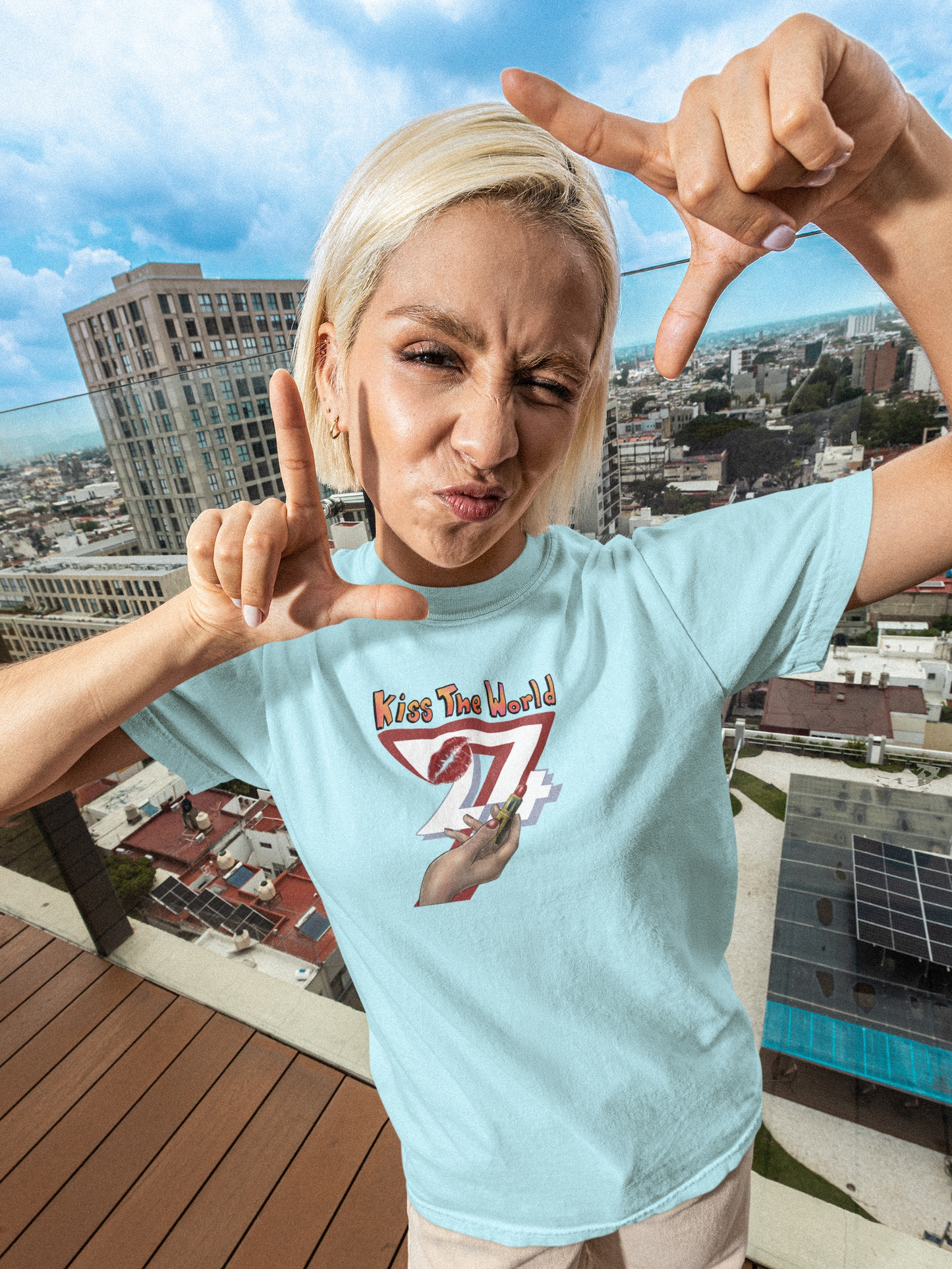 Female model looking confident wearing the light blue Kiss The World empowerment tee in a casual, sunny setting.