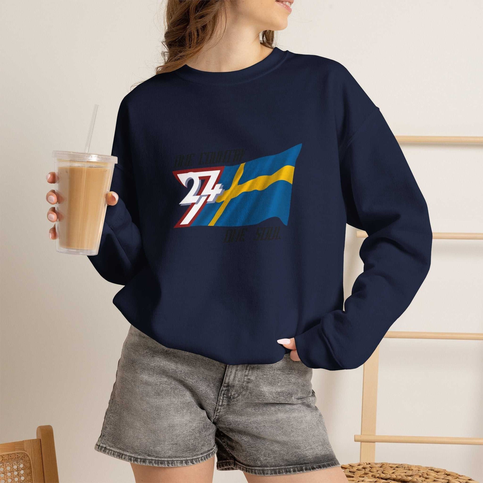 Female model styling the navy Swedish flag crewneck sweatshirt, posing casually indoors holding a coffee cup