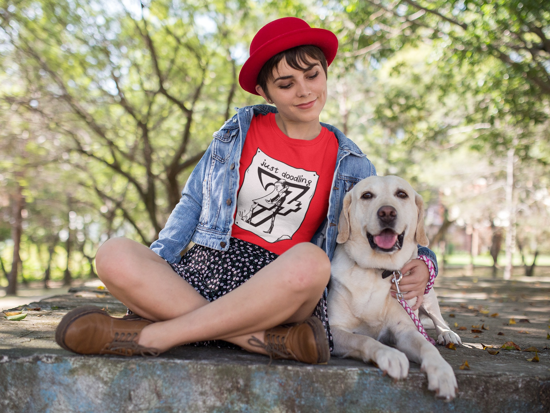 Female model wearing a red hat and the red funny dog lover graphic tee, sitting cross-legged in a sunlit park while petting a happy yellow labrador retriever.