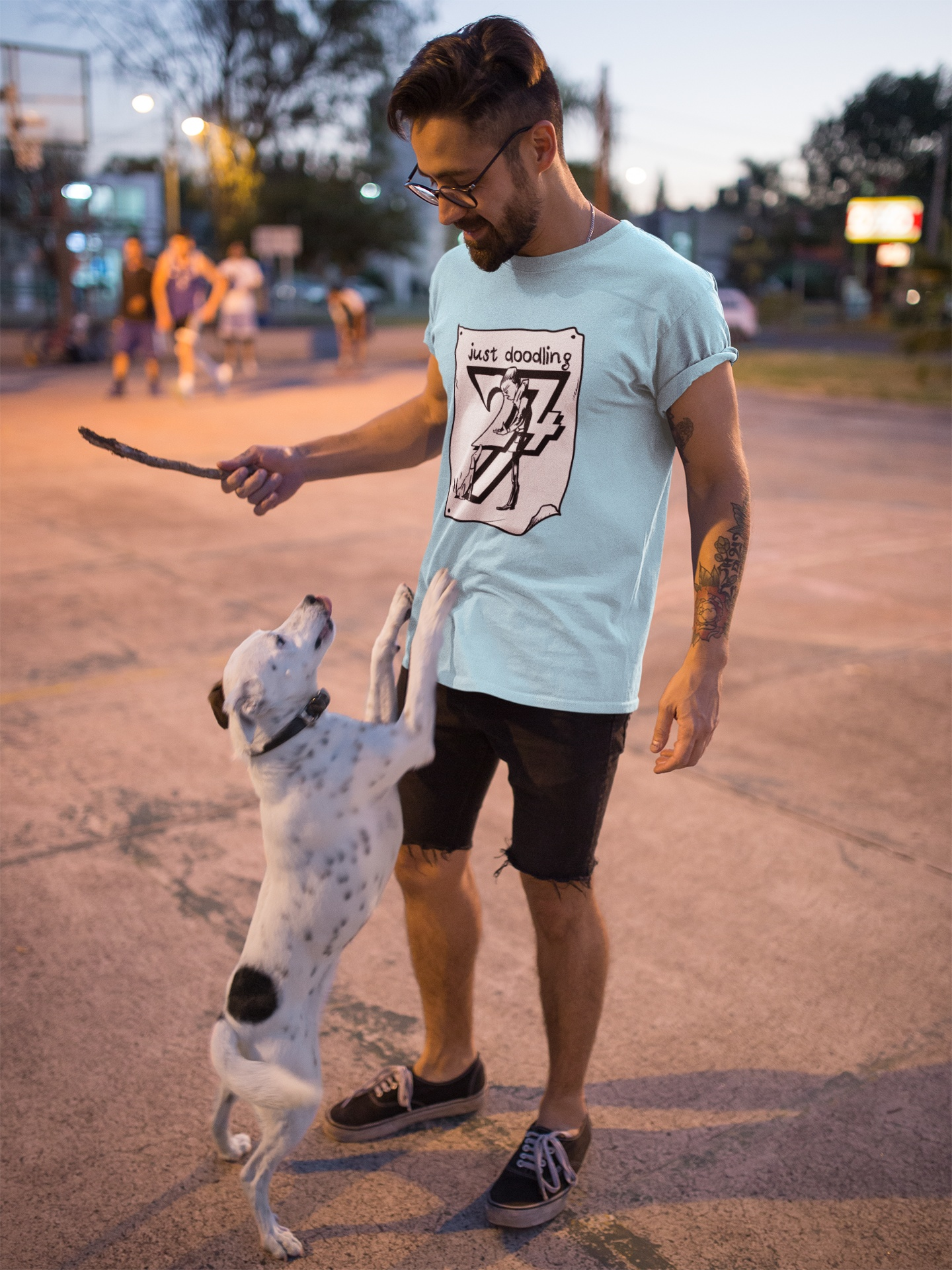 Male model wearing the light blue hand-drawn dog caricature shirt, holding a stick and playing playfully with a jumping white and black spotted dog outdoors.