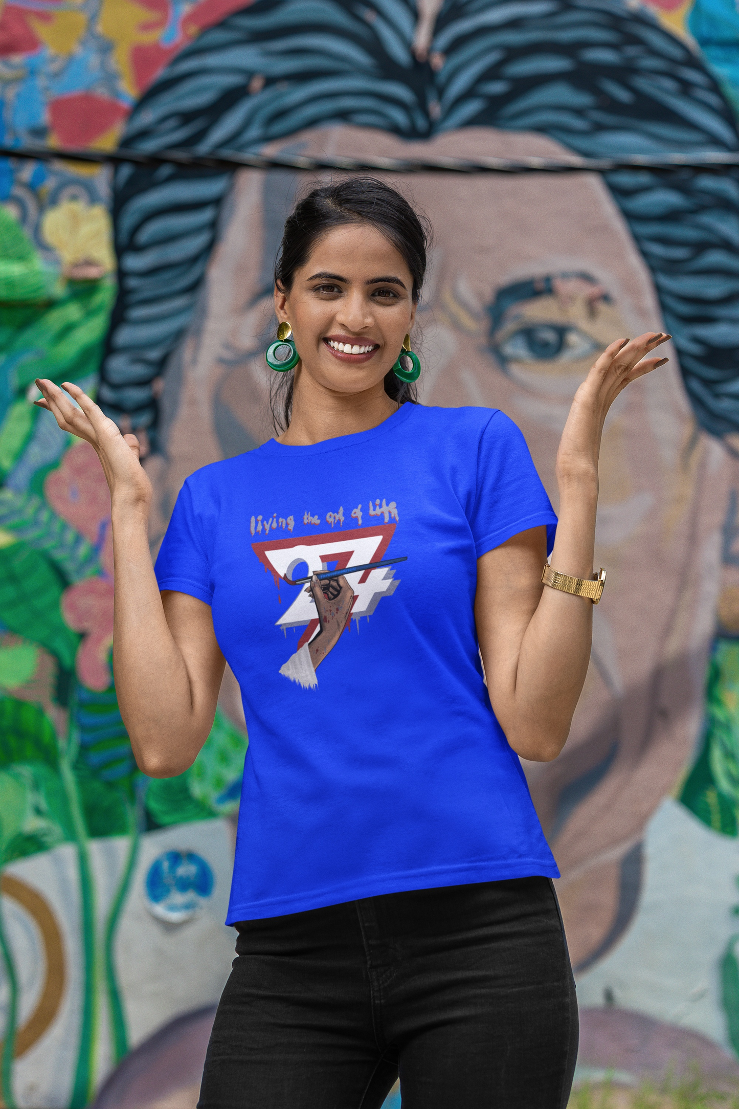 Female model wearing the royal blue female artist shirt, smiling with hands raised in a shrugging gesture against a colorful graffiti mural backdrop.